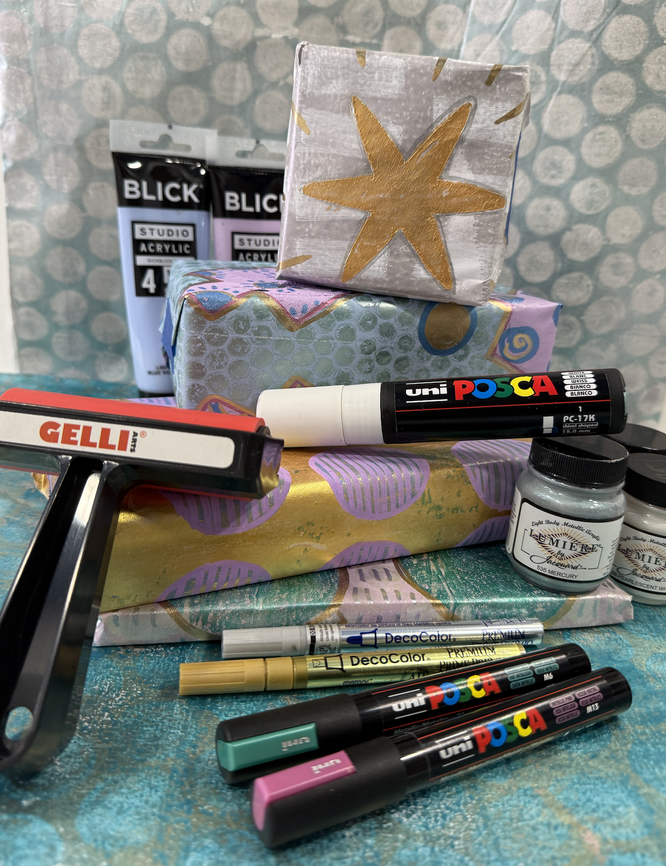 Gelli plate gift wrap supplies including gelli printing plate, acrylic paints, Posca markers, and finished custom wrapping paper with metallic gold designs