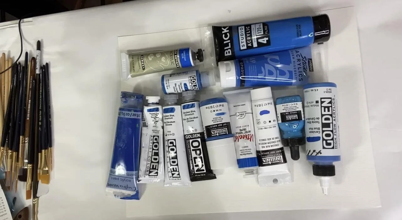 Collection of blue acrylic paint tubes and bottles from different brands lined up for testing, next to a row of paintbrushes.