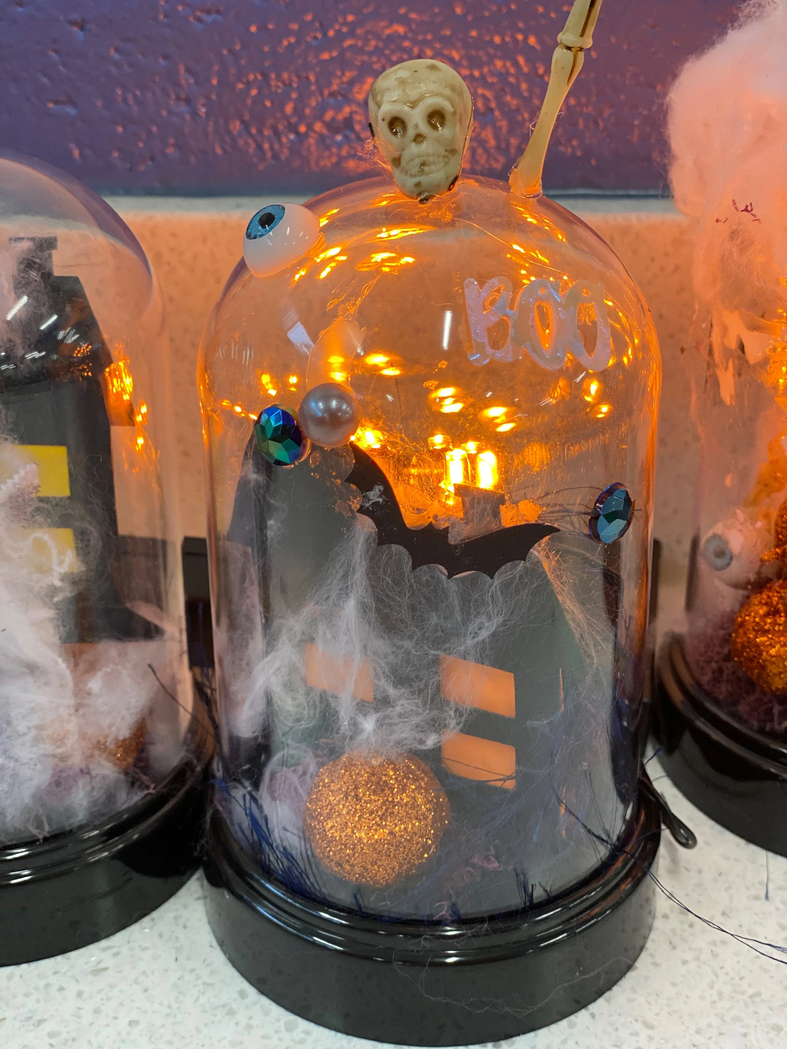 DIY Halloween cloche glowing with orange fairy lights, a haunted house, bats, pumpkin, and skeleton decorations