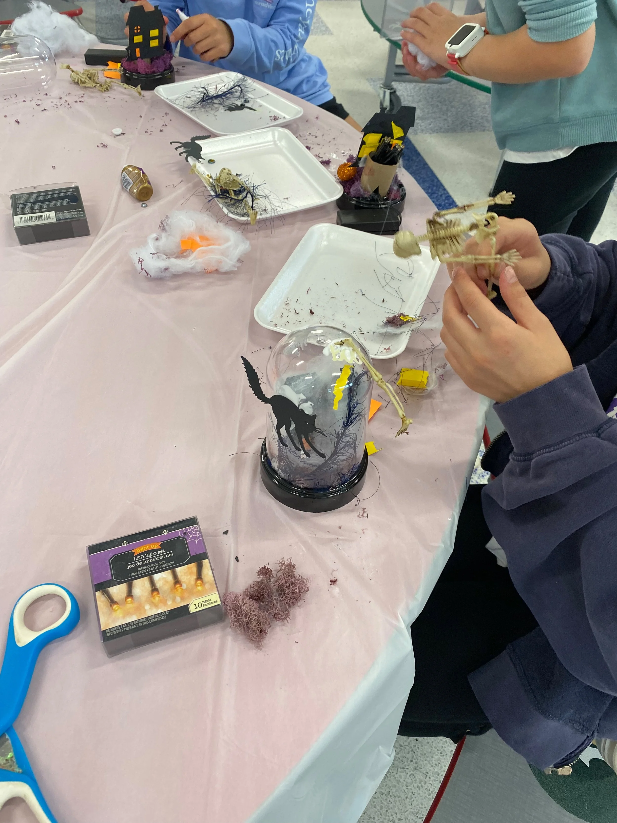 Kids making DIY Halloween cloches with miniature skeletons, haunted houses, moss, and Halloween decorations