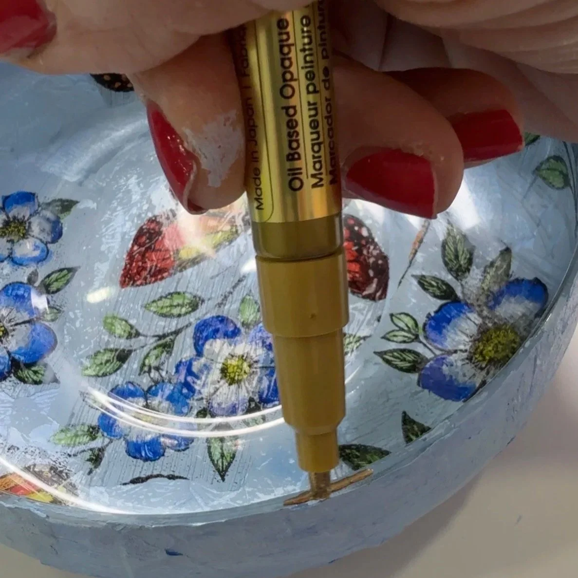 Gold oil-based paint pen adding fine edge details to a blue and orange decoupaged glass bowl