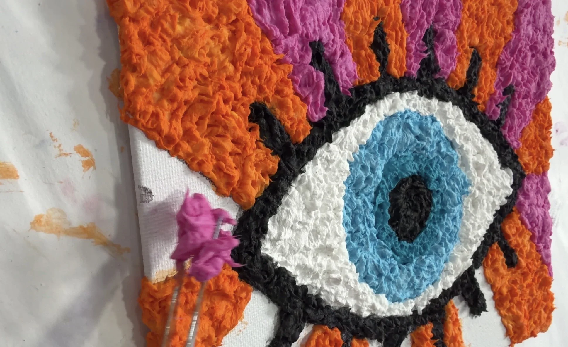 Adding pink toilet paper pulp to the orange and pink background surrounding a raised blue eye design.