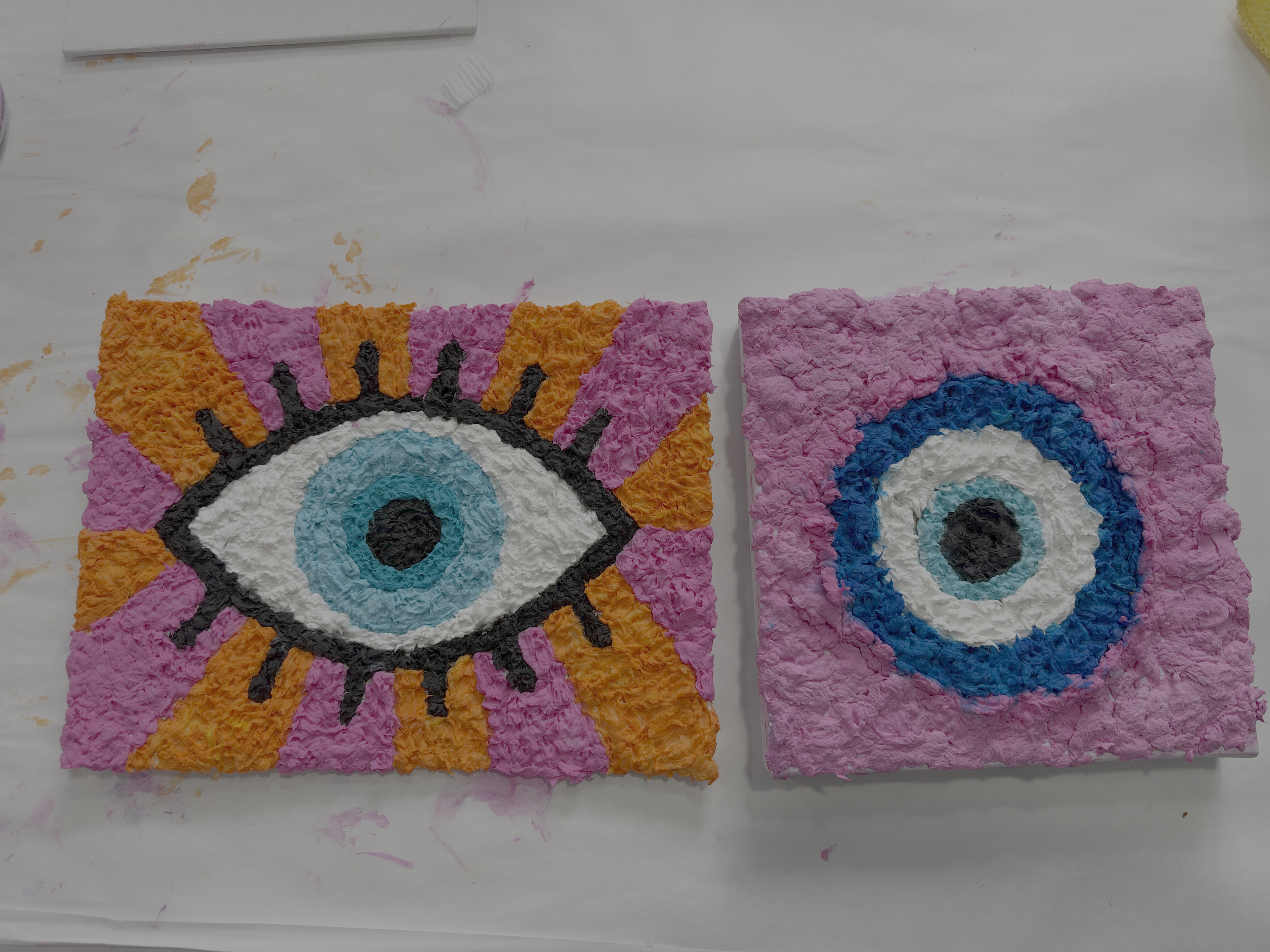Two textured toilet paper art canvases featuring a blue eye with orange and pink rays and a circular eye on pink.