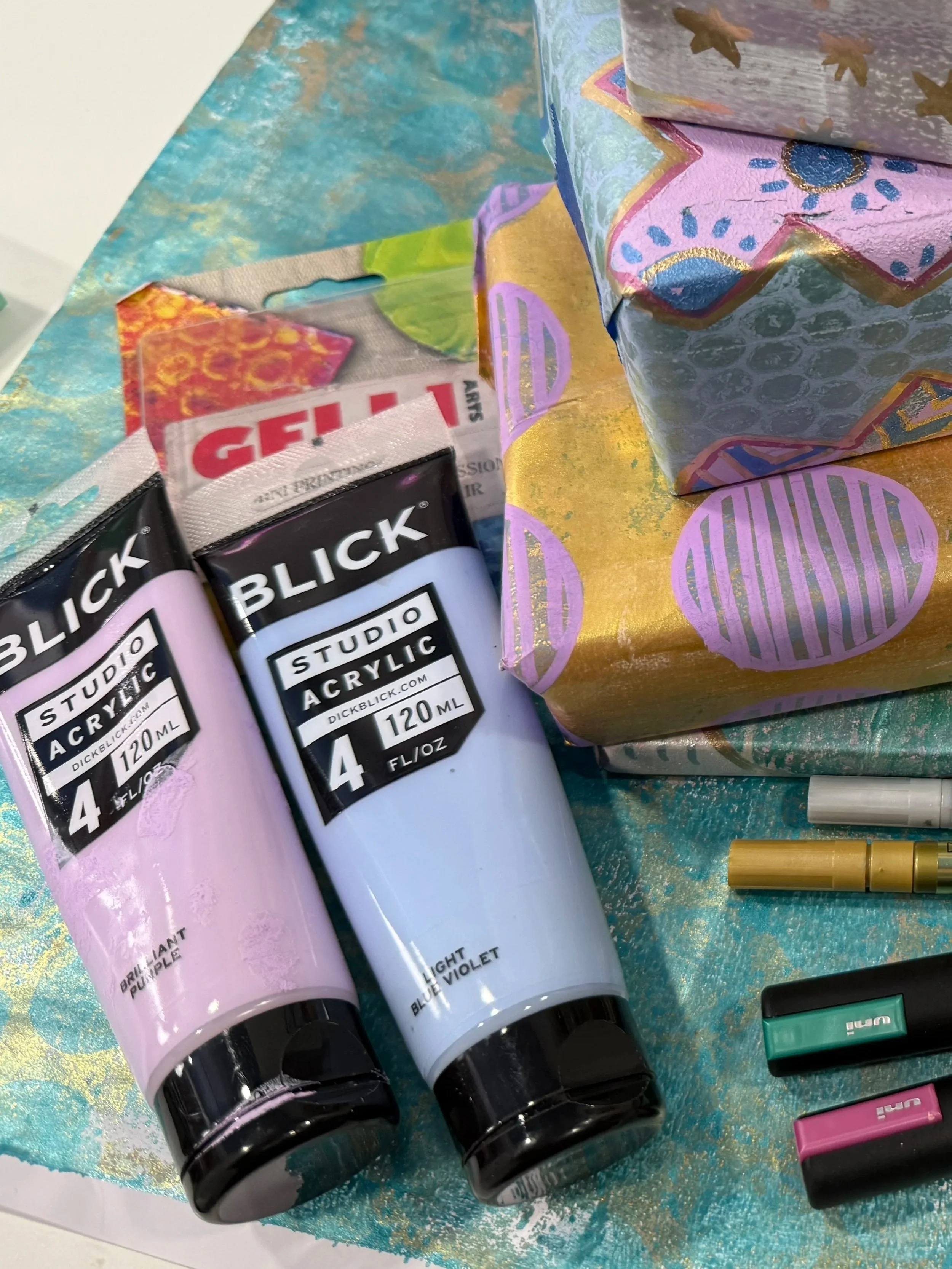 Blick Studio acrylic paints and handmade gelli plate wrapping paper samples showing bubble wrap texture and stencil designs in purple, gold, and teal