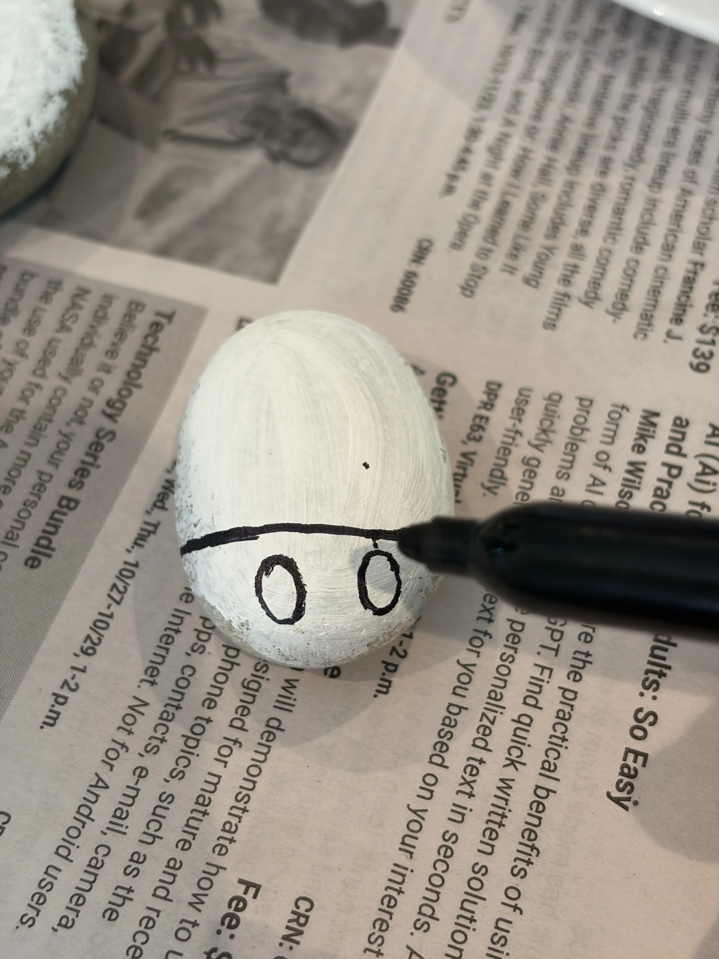 Black marker drawing the eye outlines and head line on a white-primed rock before painting a ladybug design