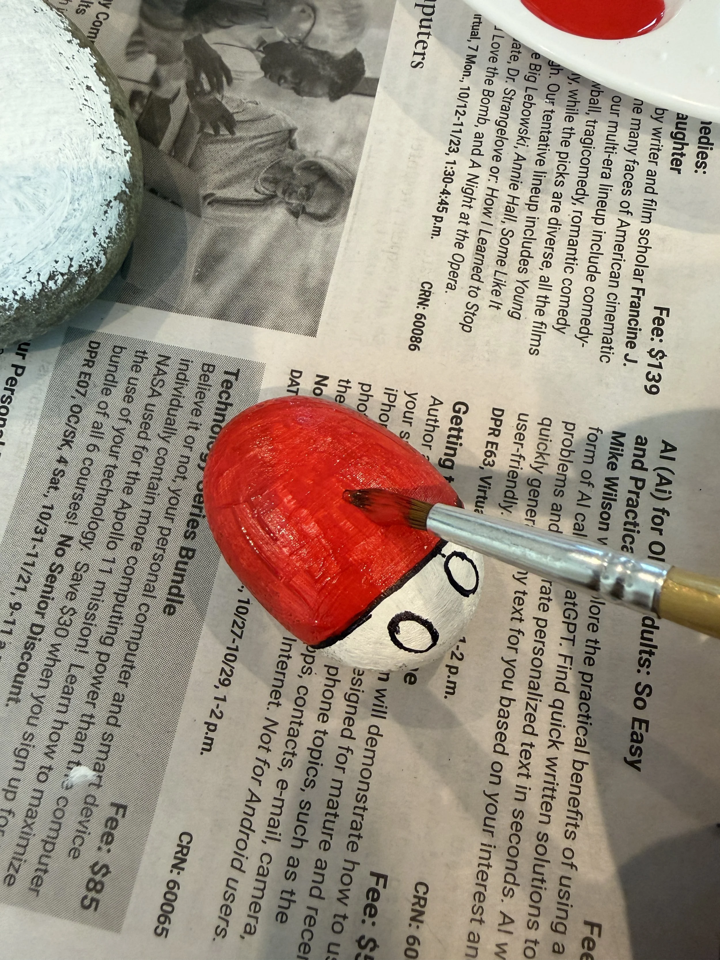 Paintbrush applying red acrylic paint over the top of a primed rock, with the black eye outlines visible along the base