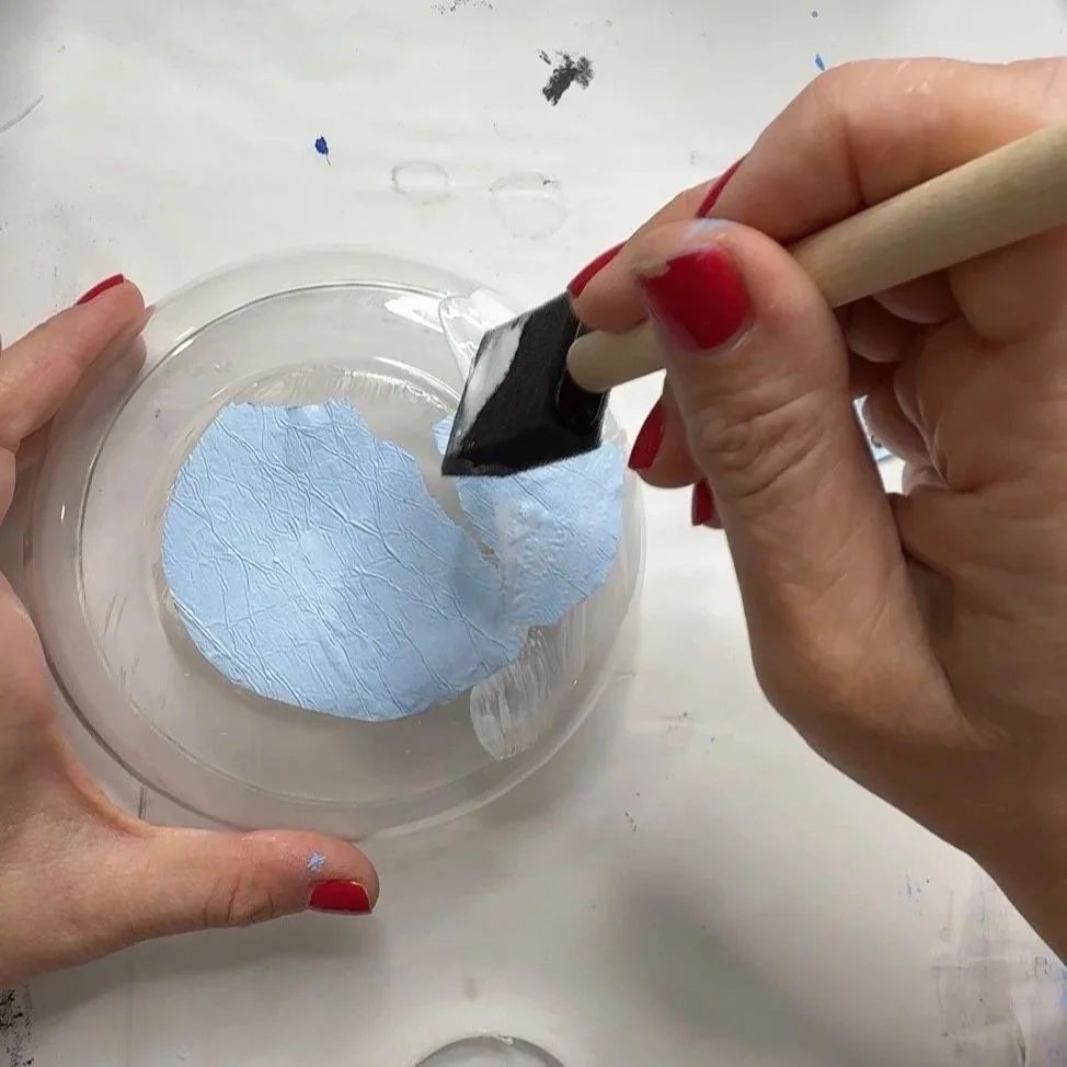 Foam brush applying Mod Podge over a blue butterfly tissue paper piece placed in a glass bowl