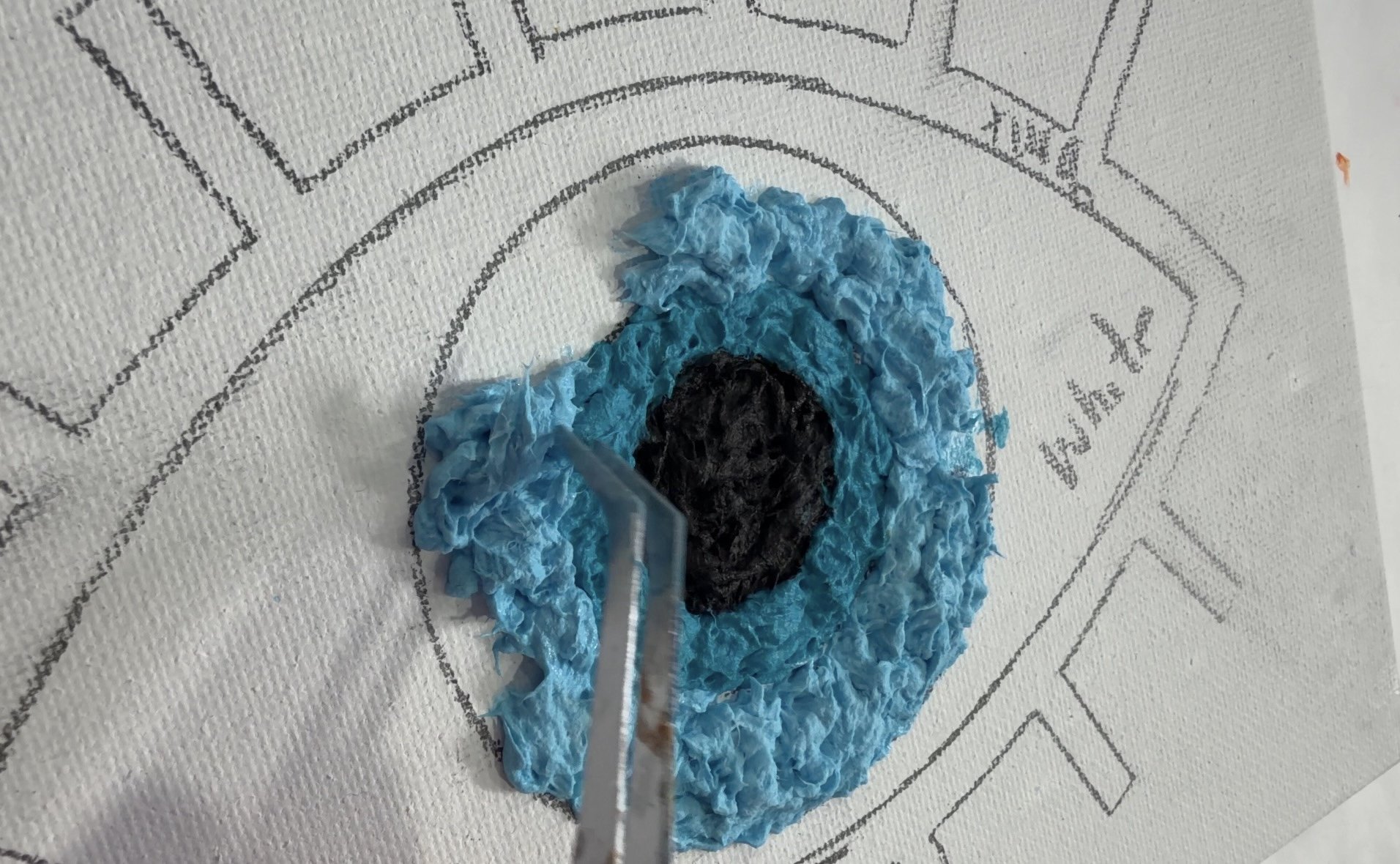 Tweezers shaping light blue toilet paper pulp around a turquoise iris to create a raised, textured eye.