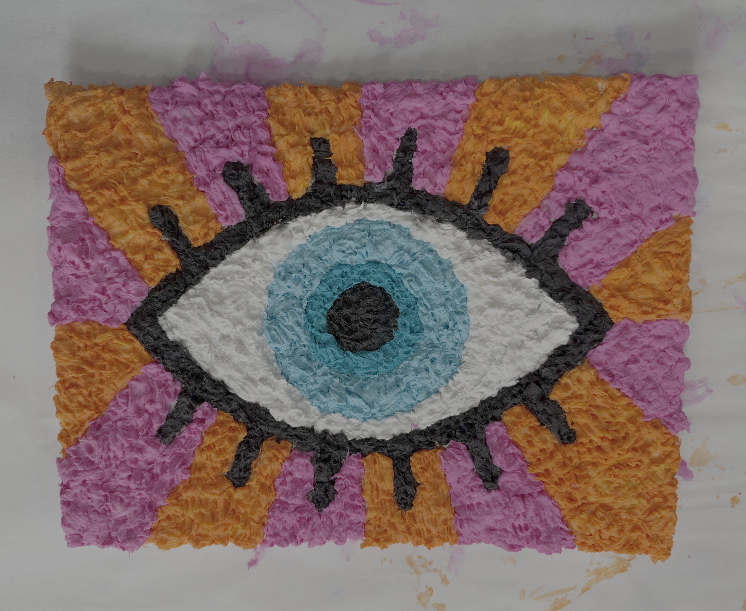 Finished toilet paper pulp art featuring a blue eye with black lashes against alternating pink and orange rays.