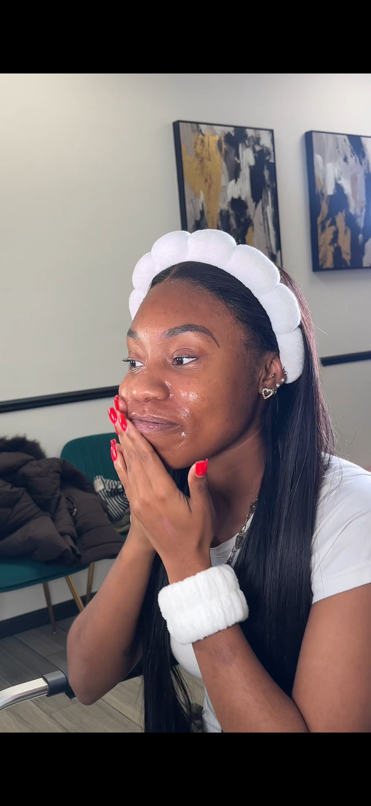 Young woman with long dark hair wearing a white headband, red nail polish, and a white sweater, sitting in a room with abstract art on the walls, touching her face with a thoughtful expression.