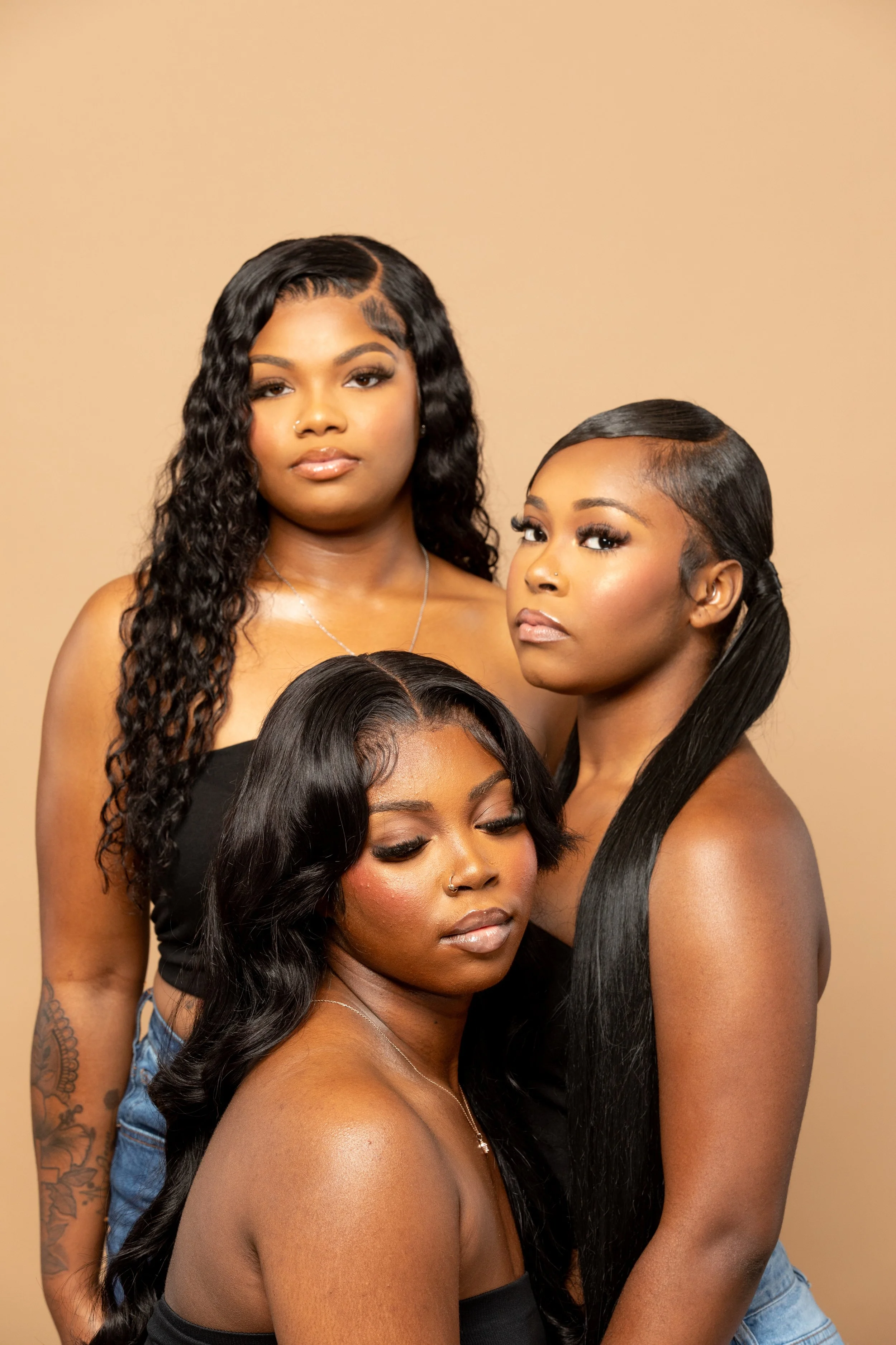 Four women with different hair styles and skin tones pose together against a plain beige background. They are dressed in black tops, with some wearing jewelry and makeup.