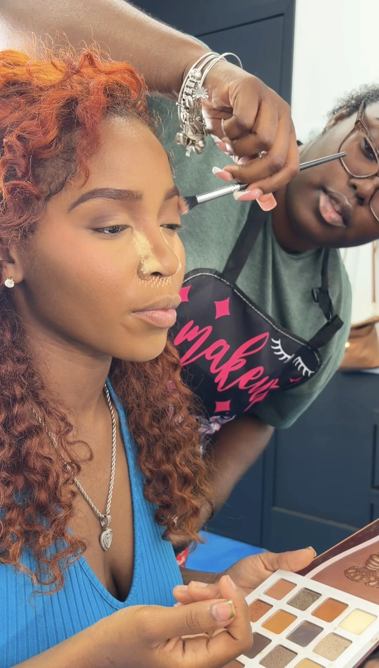 A woman with curly red hair getting her makeup done by a makeup artist using an eyeshadow palette.