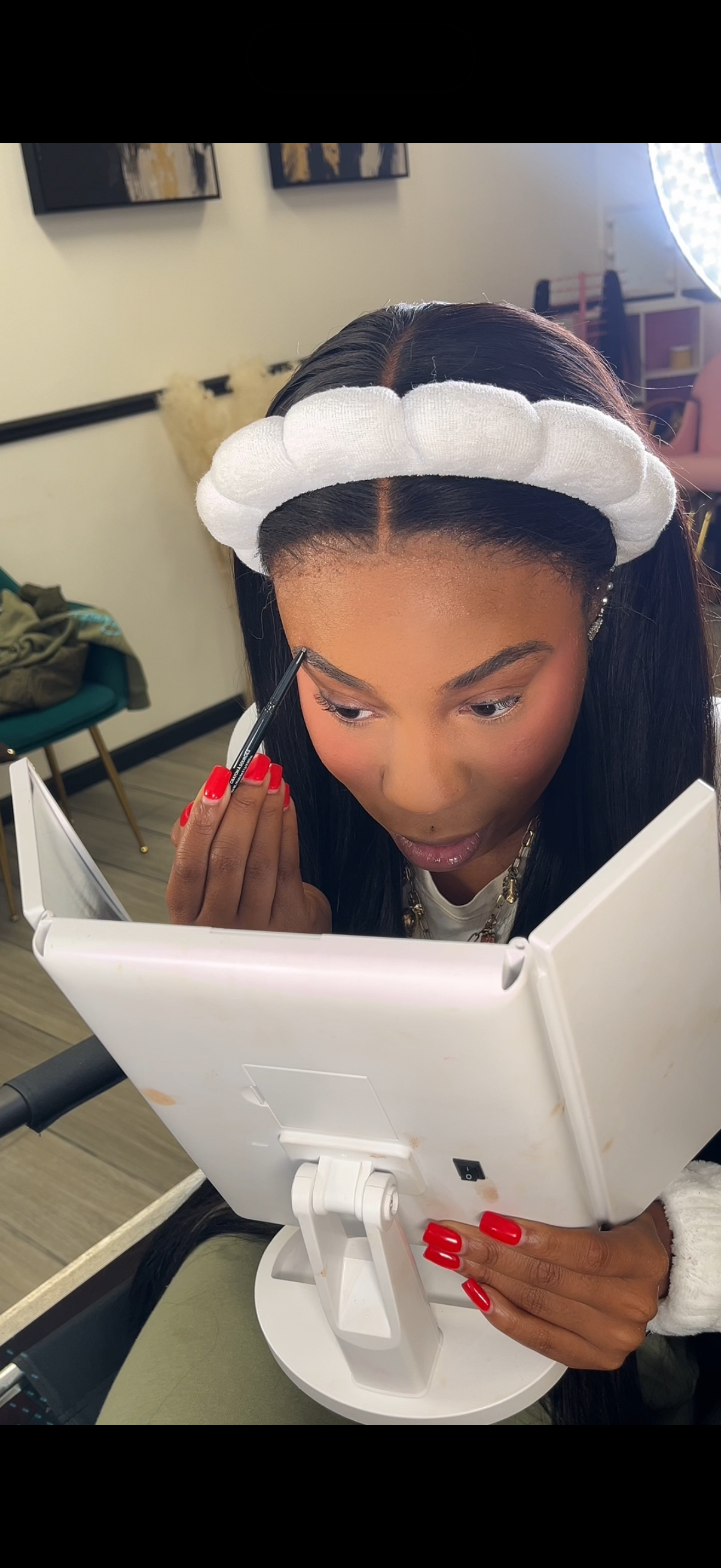 A woman with long black hair and red nails, wearing a white headband, applying makeup with an eyeliner pencil while looking in a mirror.