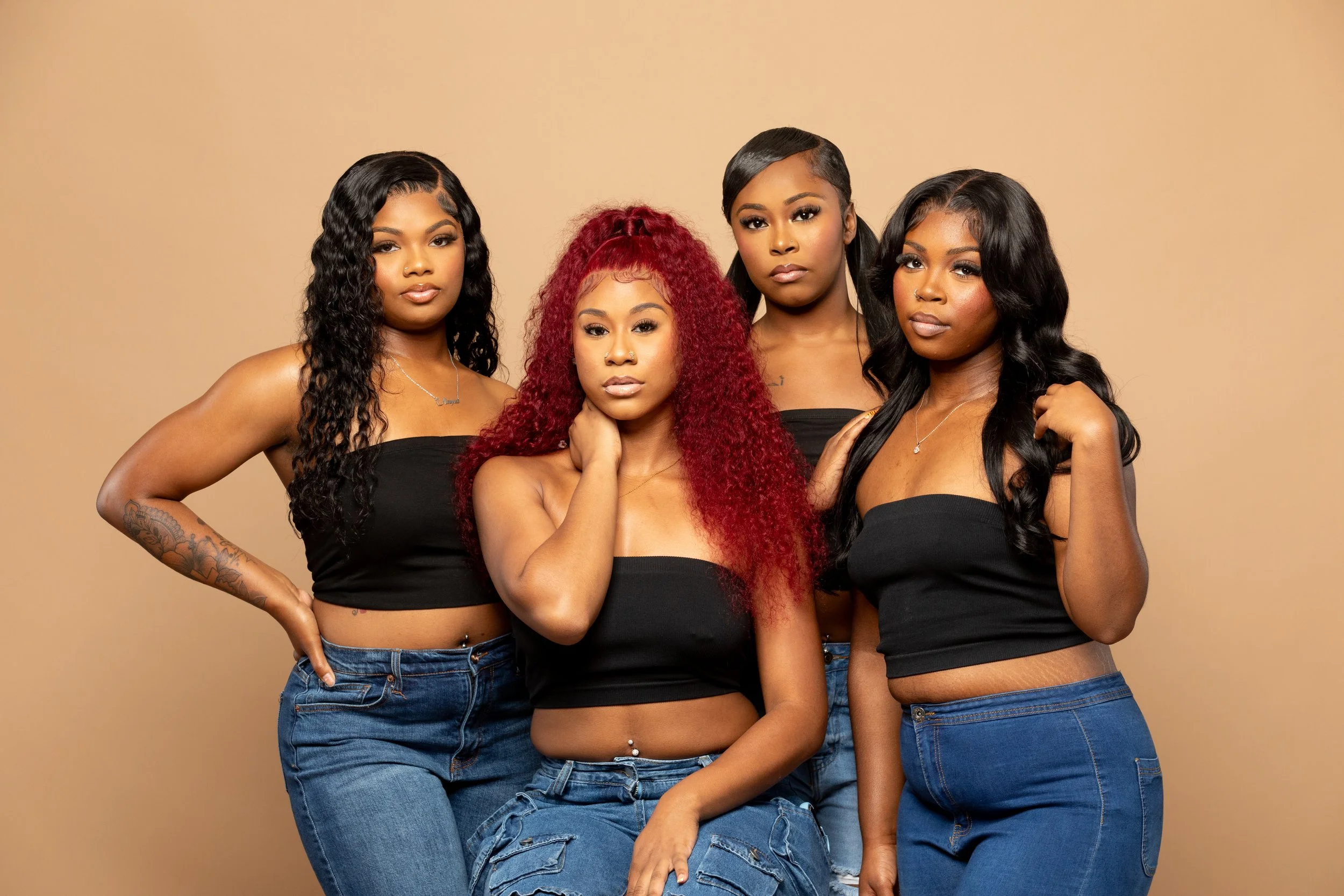 Four women posing together against a plain background, wearing black tube tops and blue jeans, with various hairstyles and accessories.