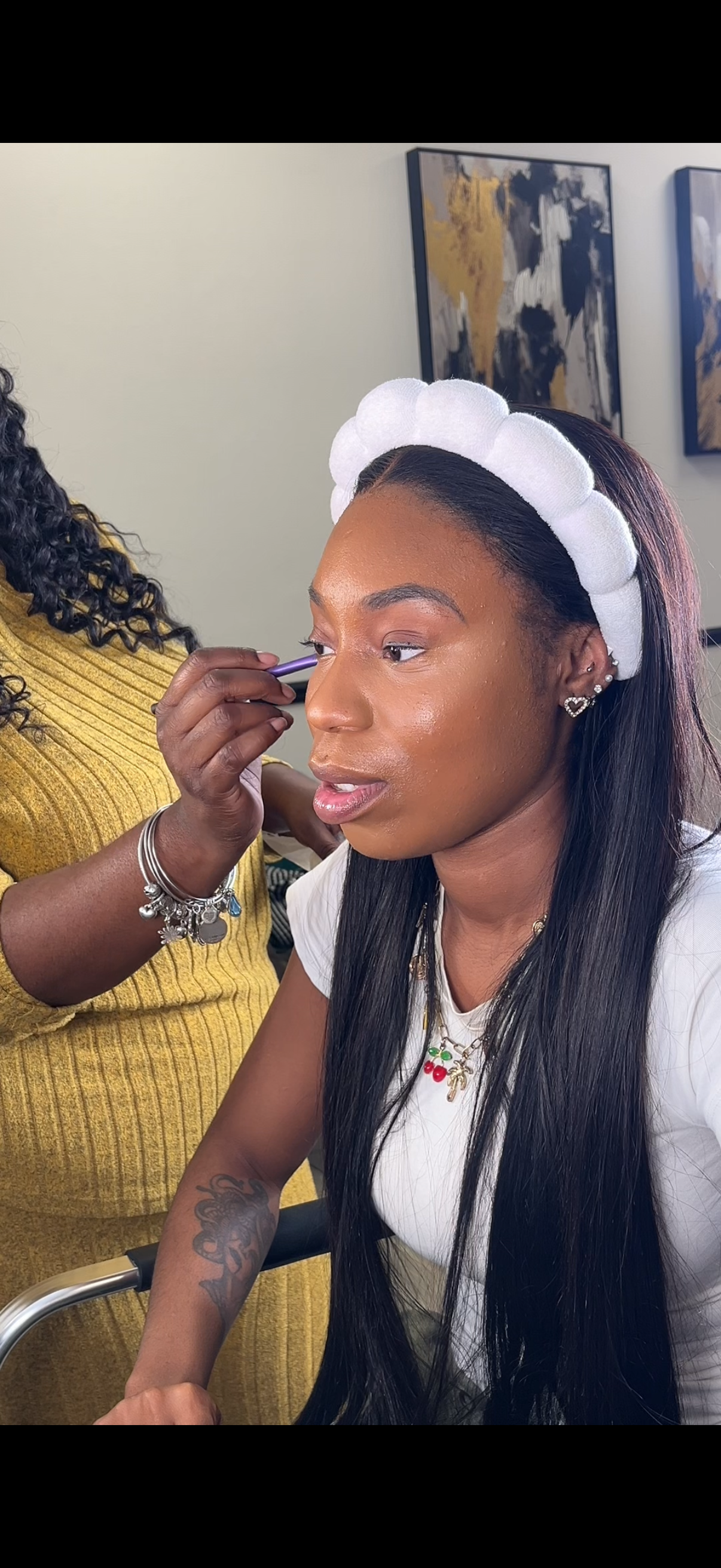 A woman with a white cloth headband having makeup applied by a makeup artist in a room with abstract world map artwork on the wall.