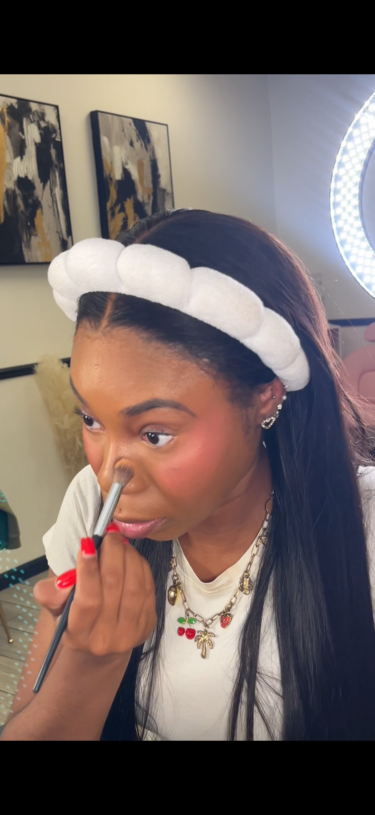 Woman applying makeup with a brush while wearing a white headband and layered necklaces, with paintings on the wall and a bright ring light in the background.