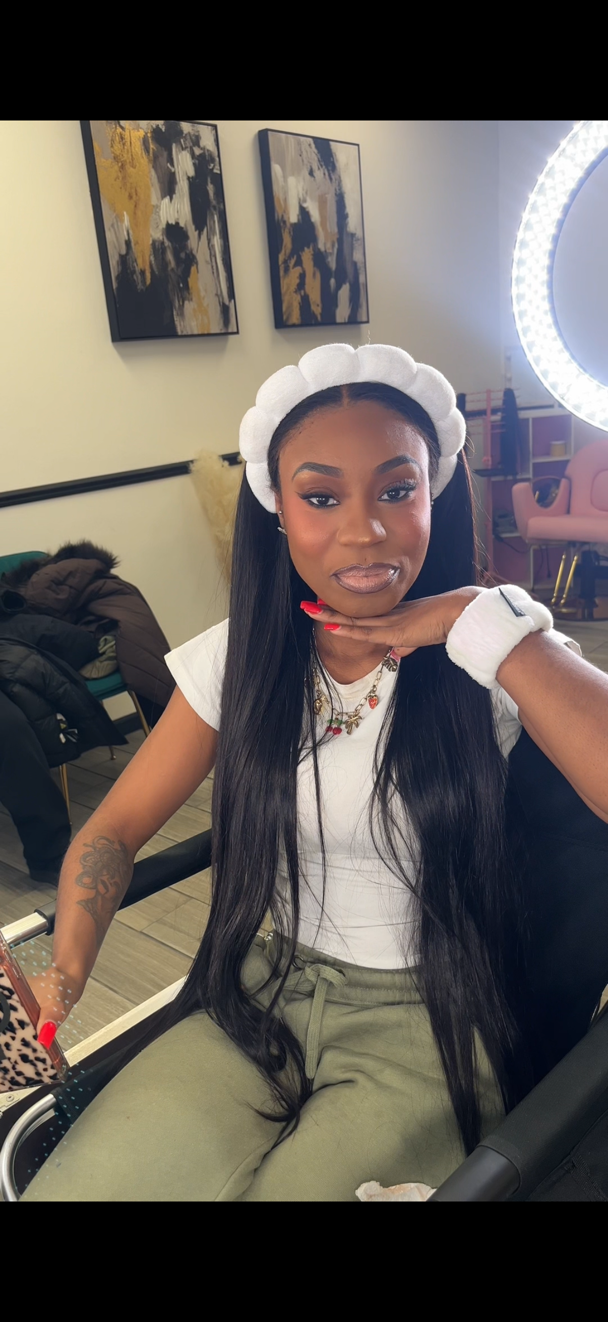 A woman with long black hair, wearing a white headband, white t-shirt, and green pants, sitting in a beauty studio. She has makeup, a colorful necklace, and a tattoo on her left arm. She is resting her chin on her left hand, with her right hand on a leopard print phone case, and looks at the camera.