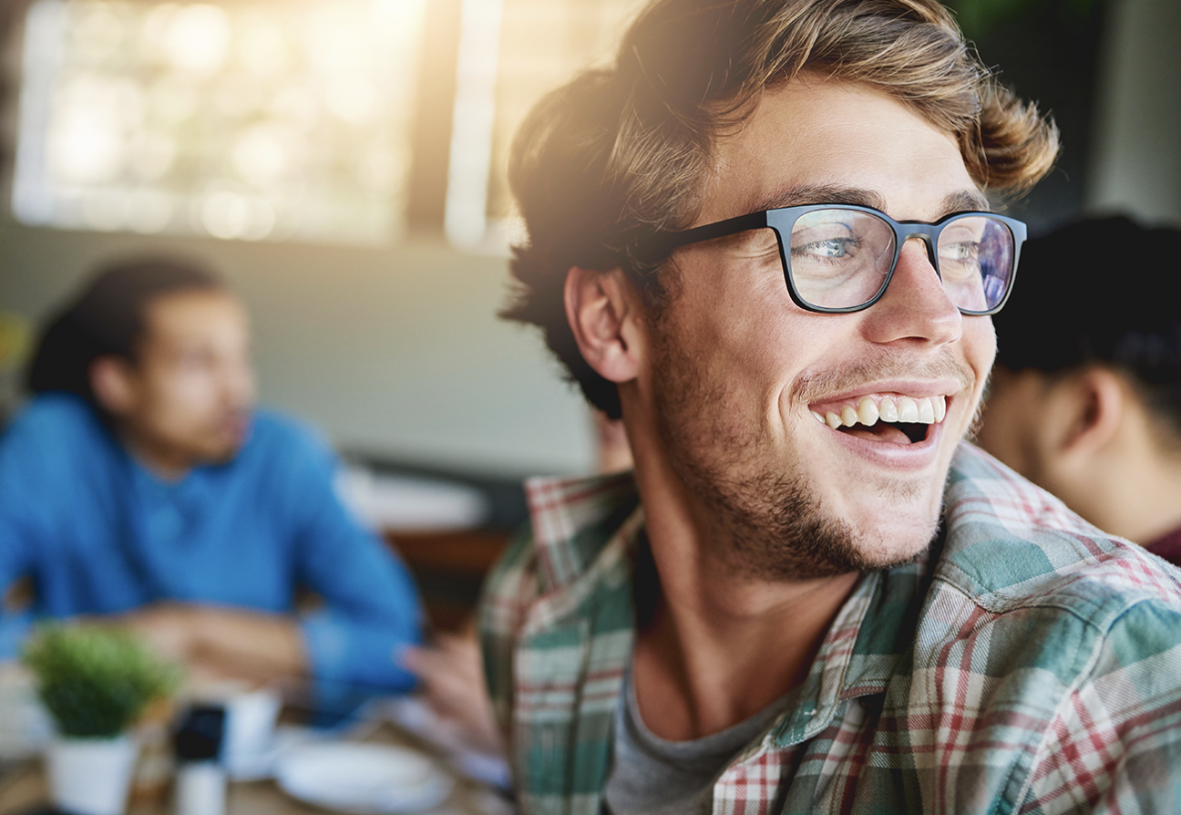 A smiling man wearing glasses and a plaid shirt is sitting indoors with blurred people in the background.