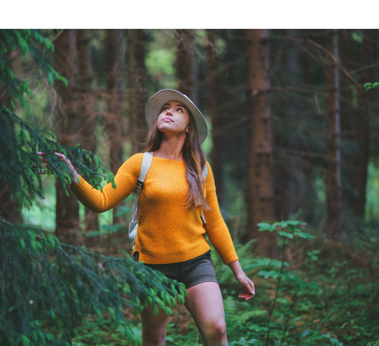 Woman wearing a yellow sweater and hat, exploring a forest.