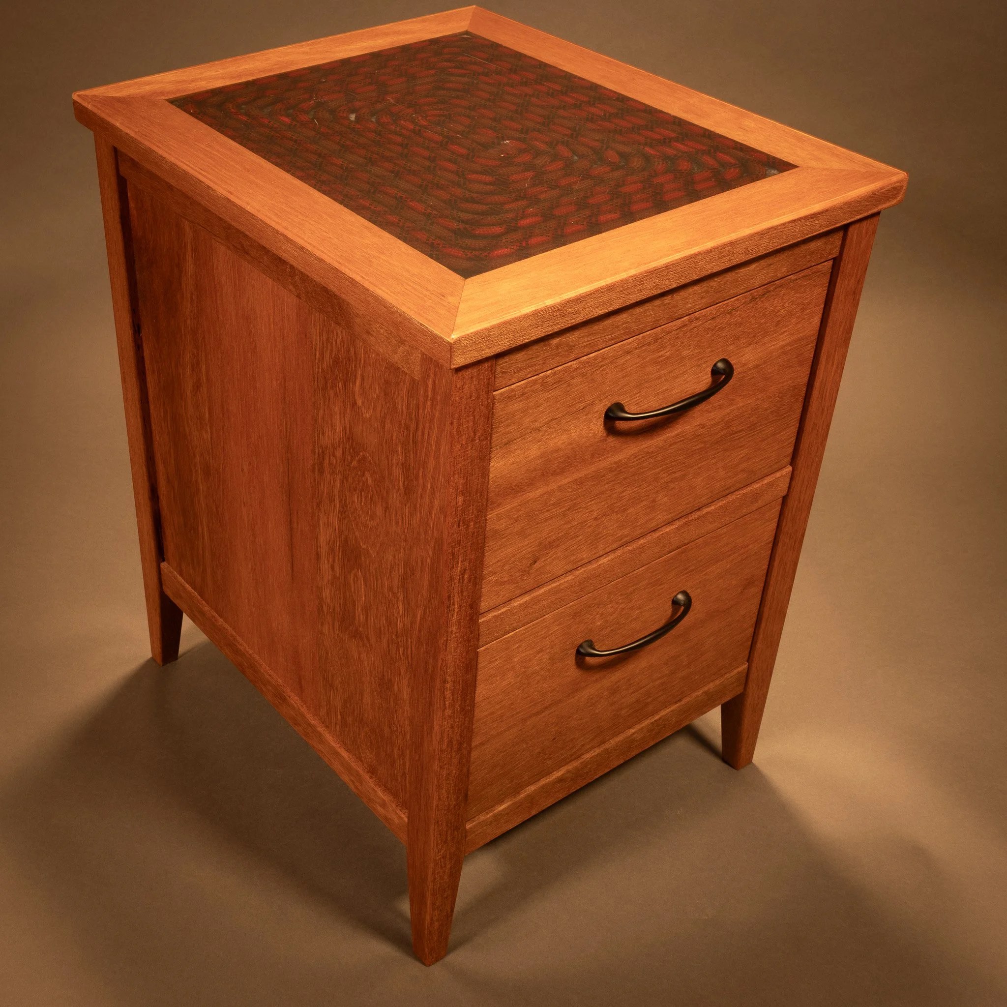 Wooden side table with a glass top, featuring a woven pattern underneath, and three drawers with black handles.