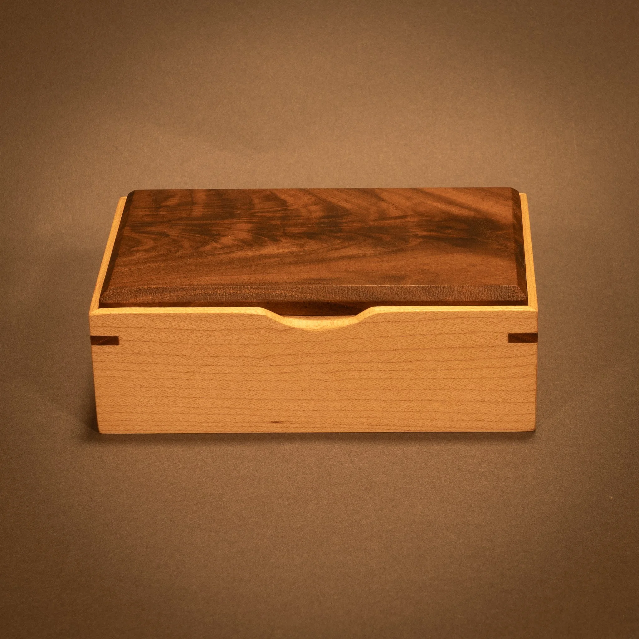 A wooden box with a rectangular shape, dark brown top surface showcasing wood grain patterns, and light-colored sides, positioned on a dark surface.