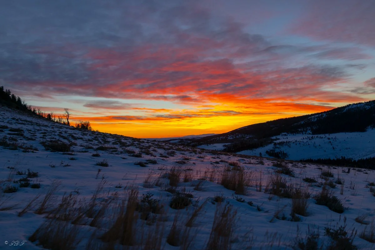 Snow-covered hills at sunset with colorful orange, pink, and purple sky and scattered clouds.