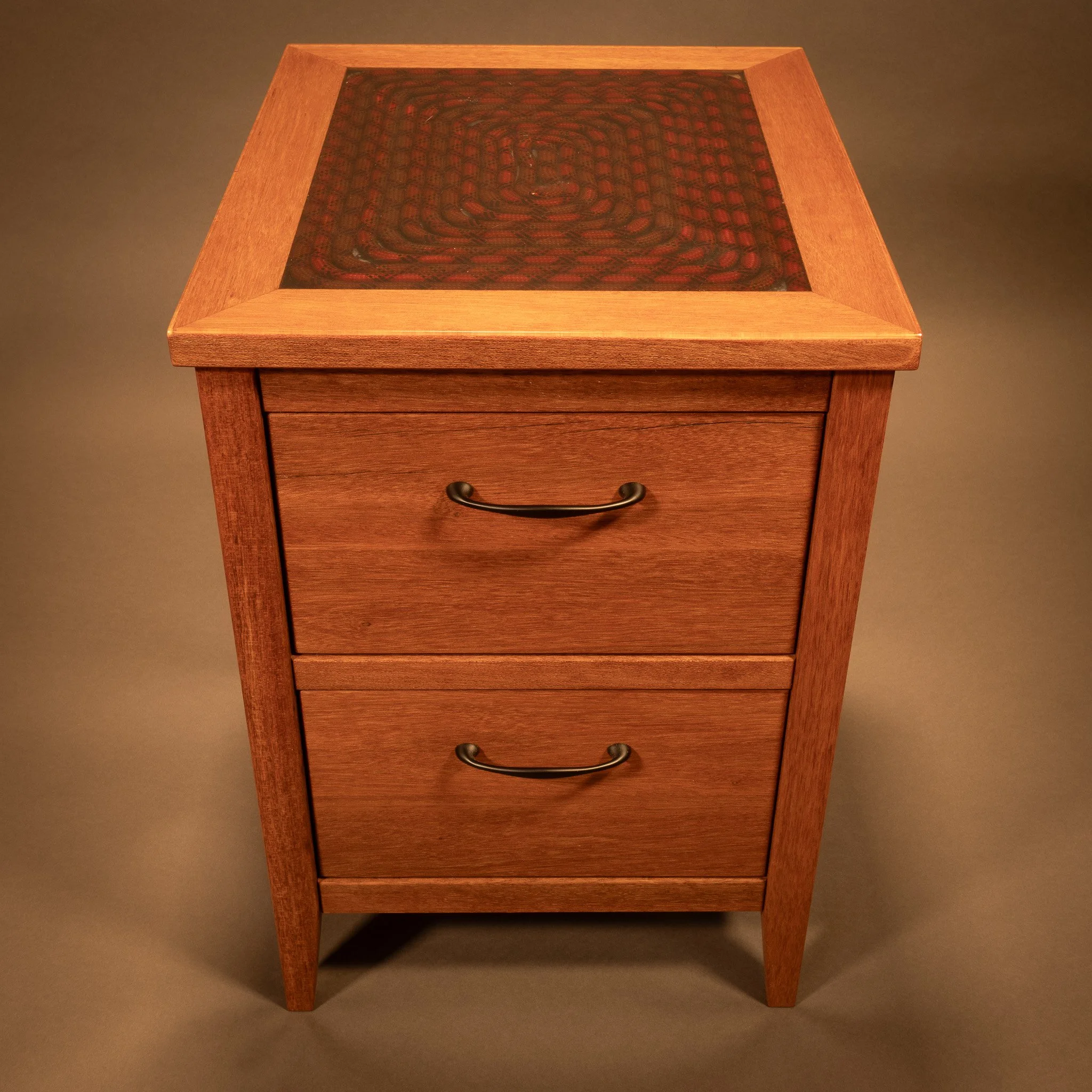 A wooden side table with a glass top, displaying a dark patterned design underneath, positioned against a plain, neutral-colored background.