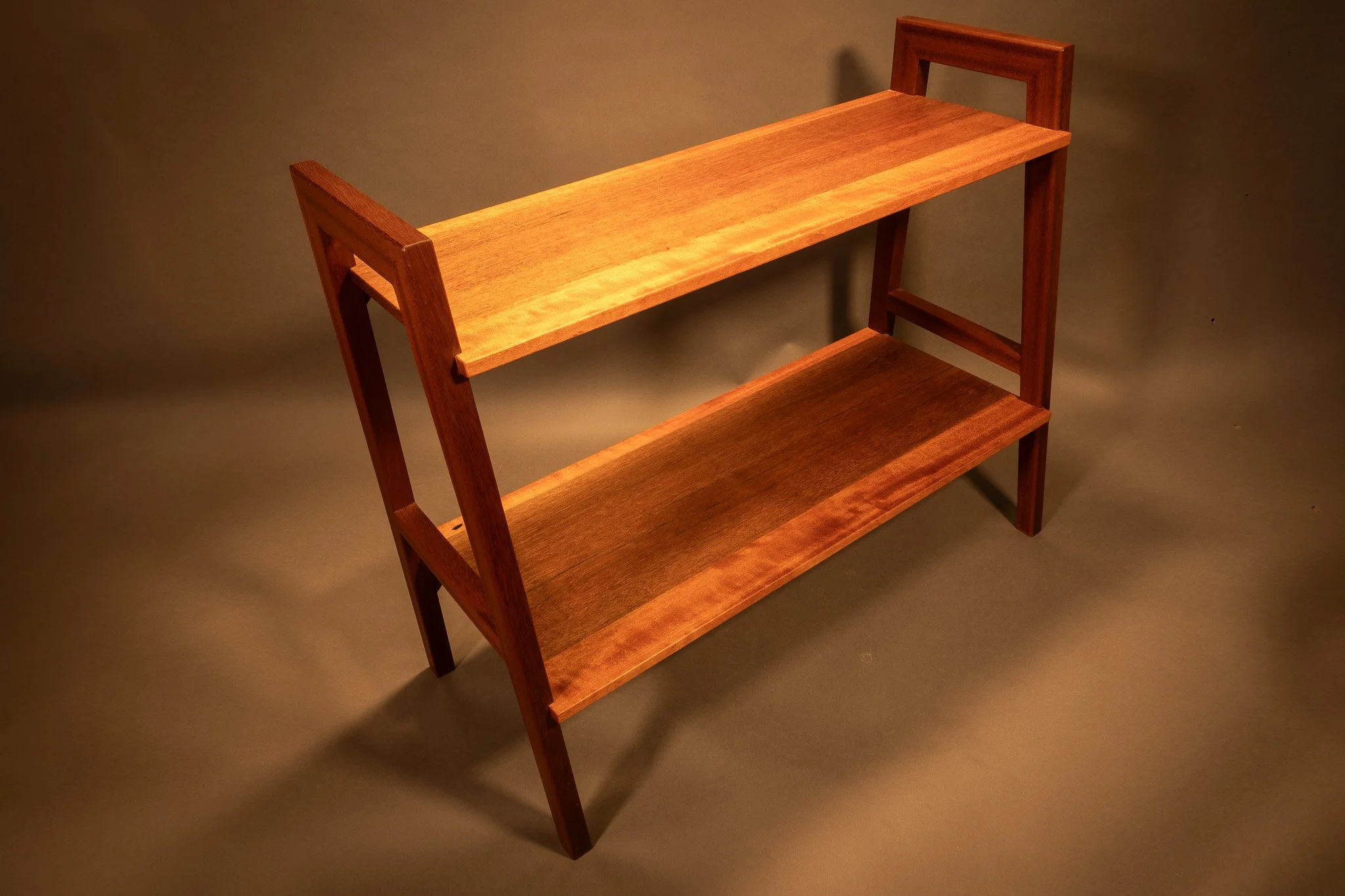 A wooden multi-tiered shelf or stand with three shelves, made of reddish-brown wood, against a plain neutral background.
