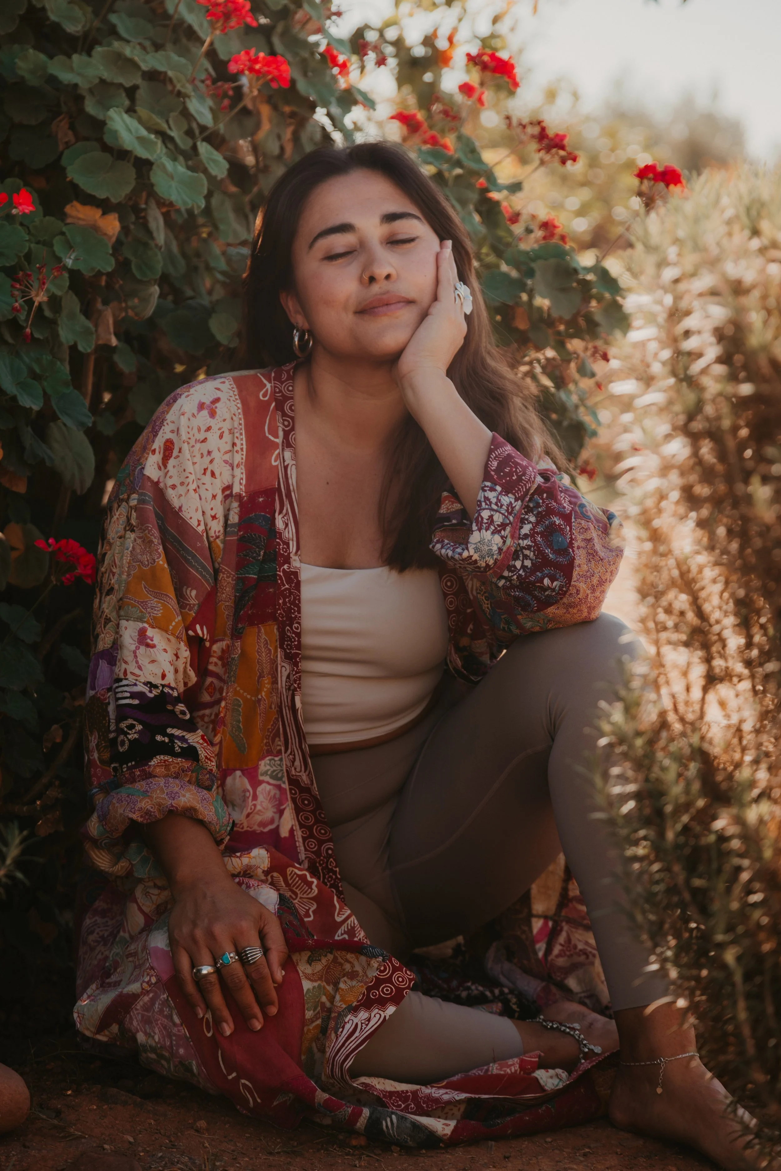 A woman sitting outdoors with closed eyes, wearing a colorful patterned shawl and beige leggings, surrounded by green foliage and red flowers.
