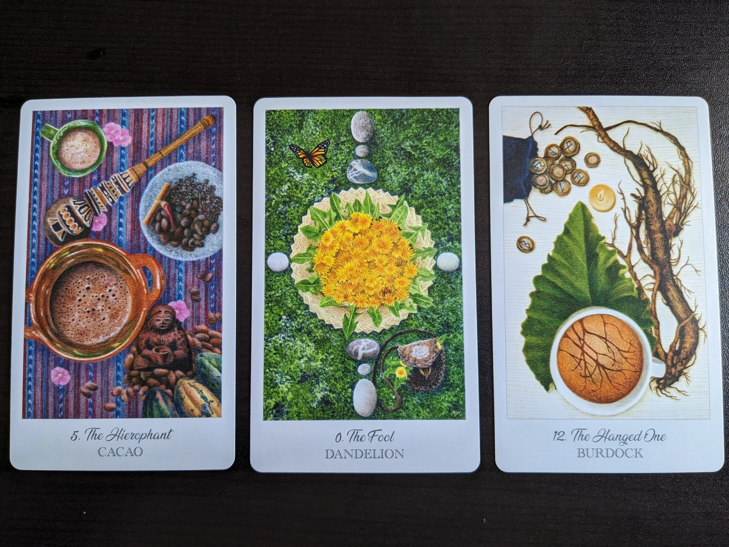 Three tarot cards laid out on a dark surface. The first card, labeled 'The Hierophant,' shows a cup of cacao, a bowl of cocoa beans, and a wooden musical instrument. The second card, 'The Fool,' features a dandelion blossom with surrounding stones, leaves, and a butterfly. The third card, 'The Hanged One,' depicts a cup of hot beverage with roots, a leaf, and seed pods.