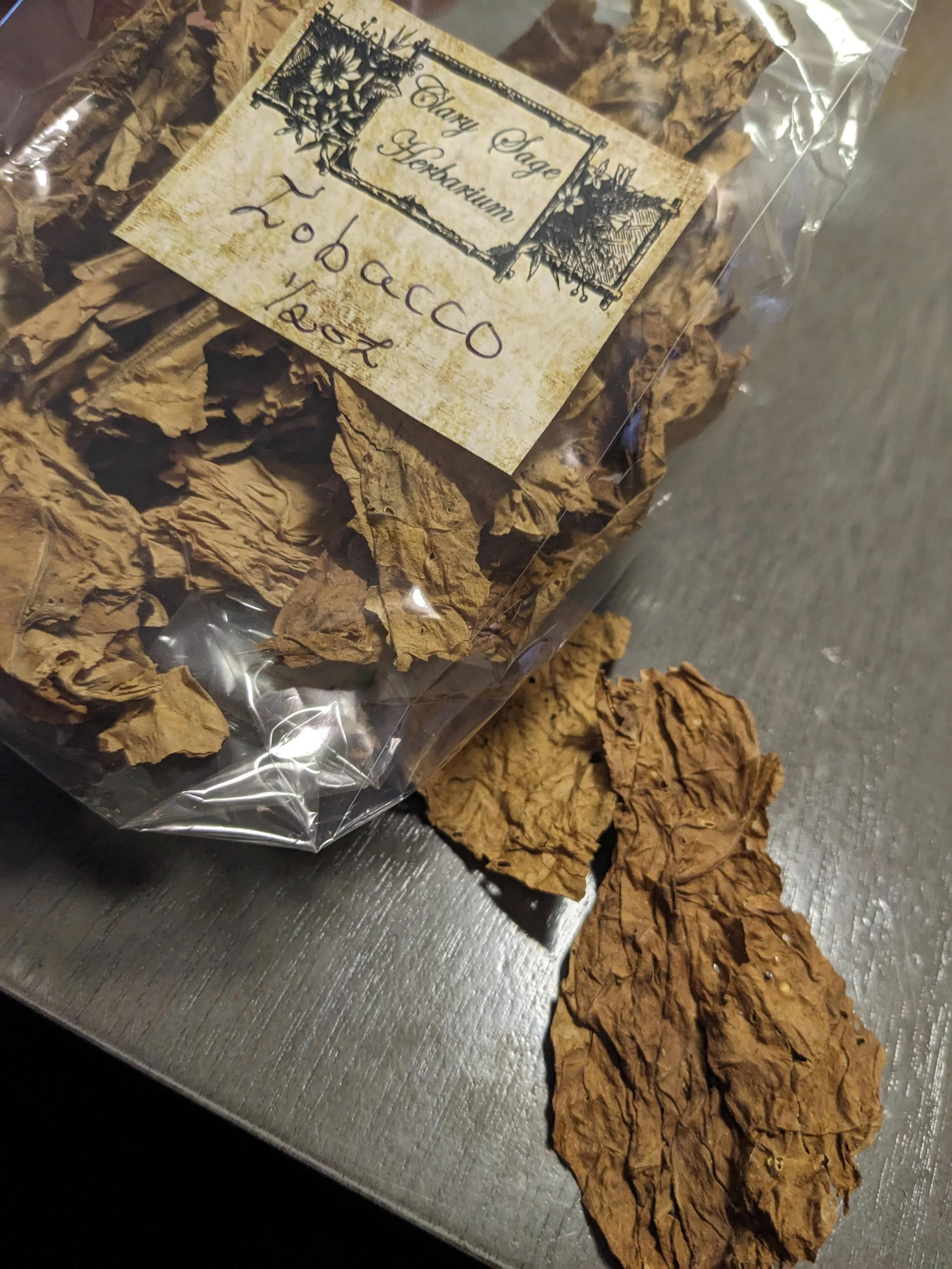 A clear plastic bag of dried tobacco leaves with a label that reads 'Hay Sage Herbarium Tobacco 12oz', placed on a dark surface with some tobacco leaves spilled out.