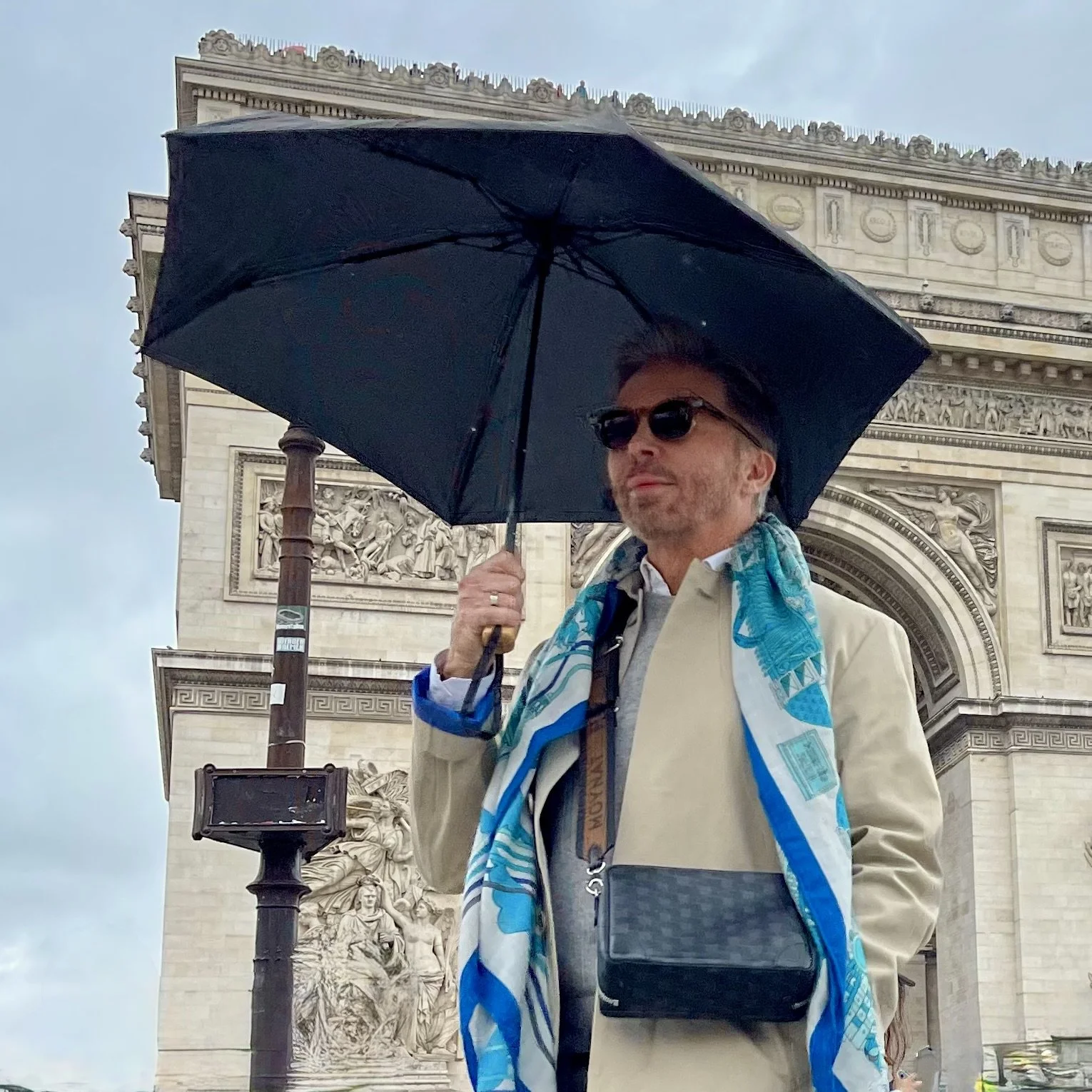 Man in a beige trench coat holding a black umbrella in front of the Arc de Triomphe on a rainy day, wearing sunglasses, a blue patterned scarf, and a crossbody bag.