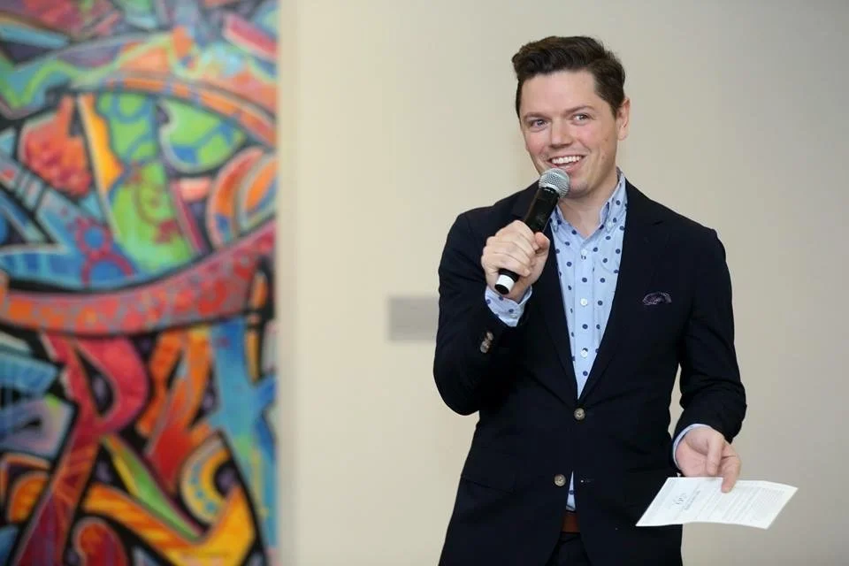 David Peck speaking at an event, holding a microphone and notes, in front of a colorful abstract painting