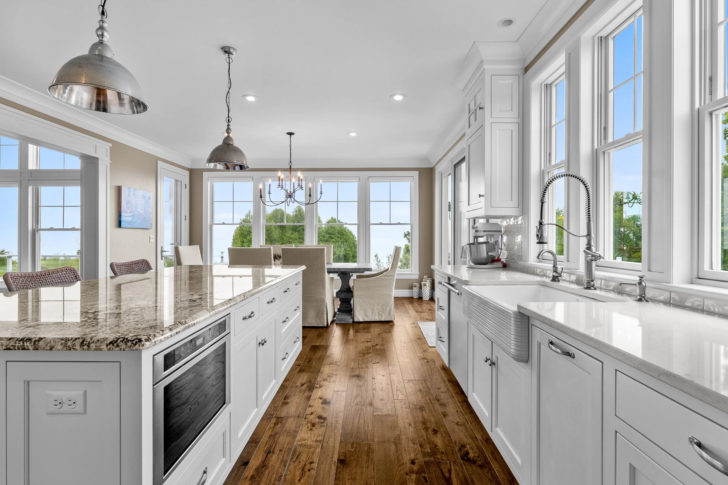 A bright, spacious kitchen with white cabinets, a large island with a granite countertop, and hardwood floors. There are multiple windows with views of greenery outside, and a dining area with a table and chairs in the background. Pendant lights hang
