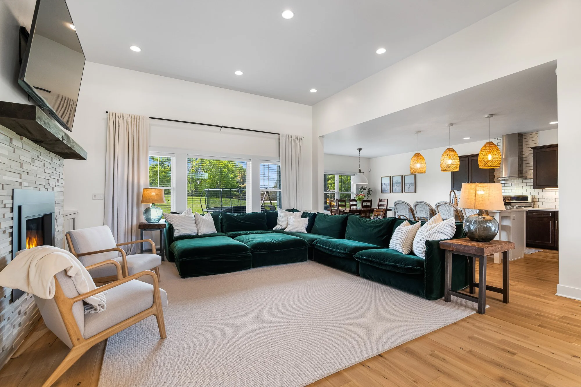 Spacious living room with a green velvet sectional sofa, armchair, fireplace, and large windows, connected to a dining area and kitchen with hanging pendant lights.