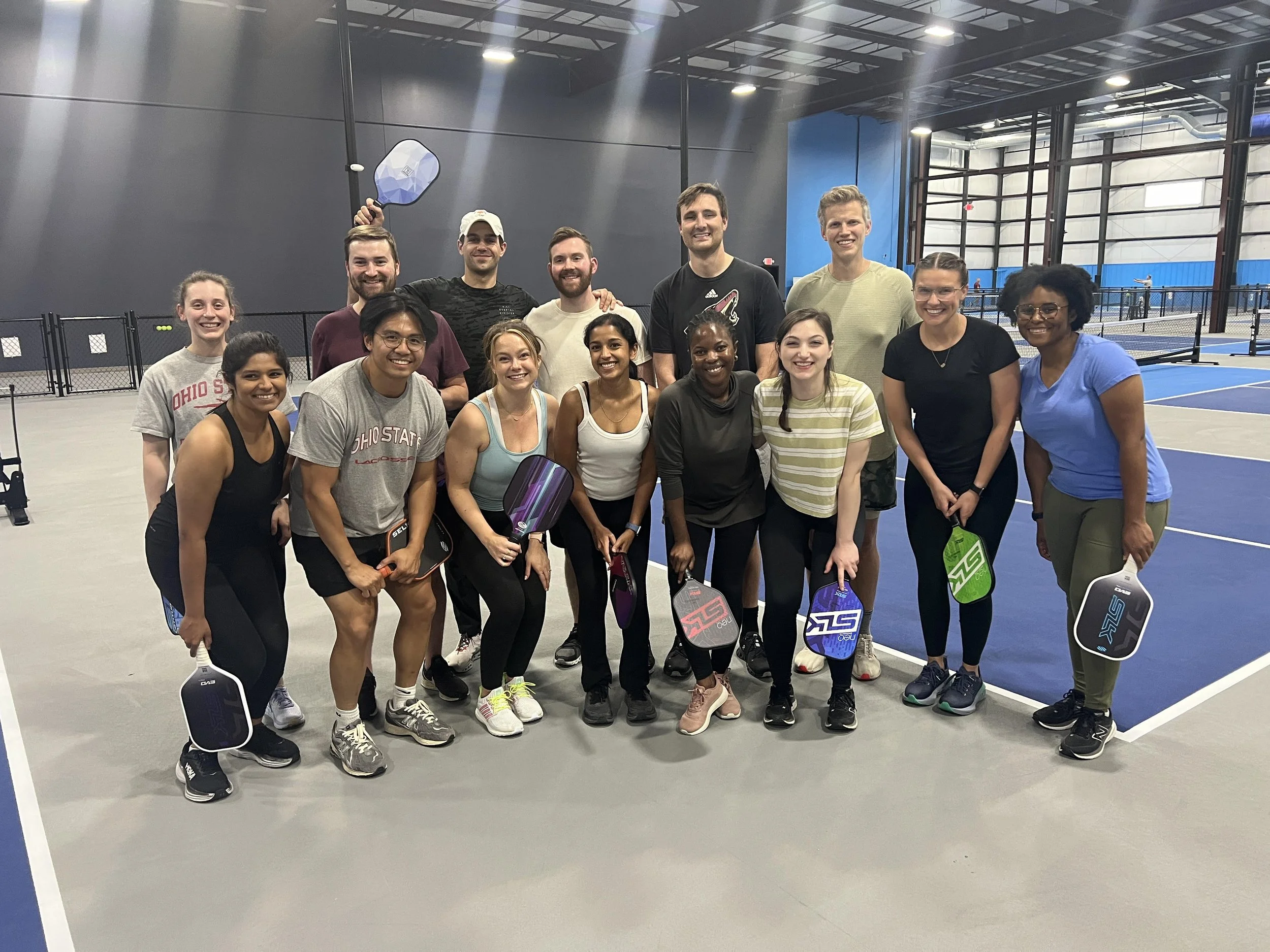 A group of fifteen people standing on a tennis court indoors, holding pickleball paddles and smiling for the camera.
