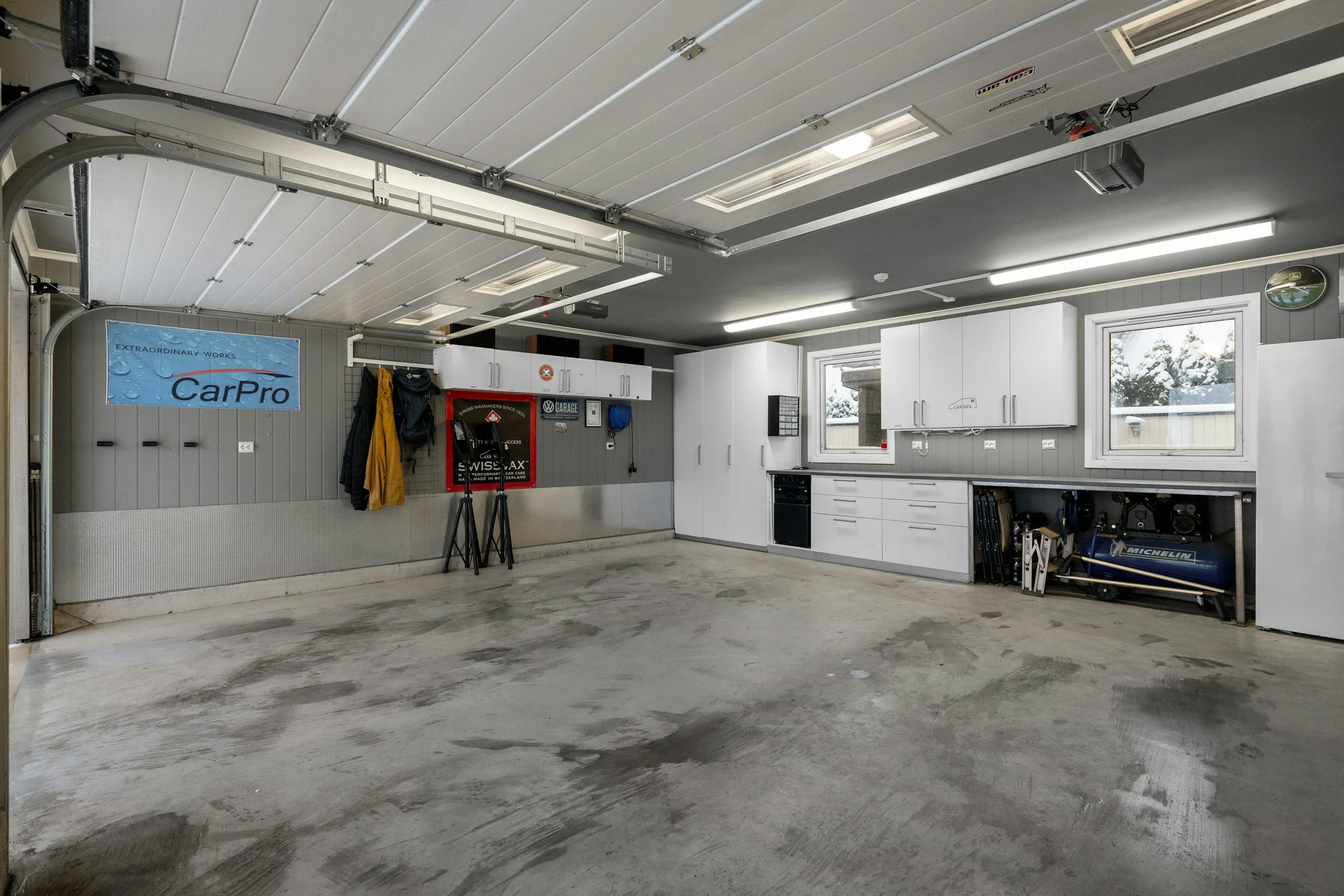 Empty garage with white cabinetry, two windows, epoxy or polyurea floor, and various tools and equipment stored along the walls.