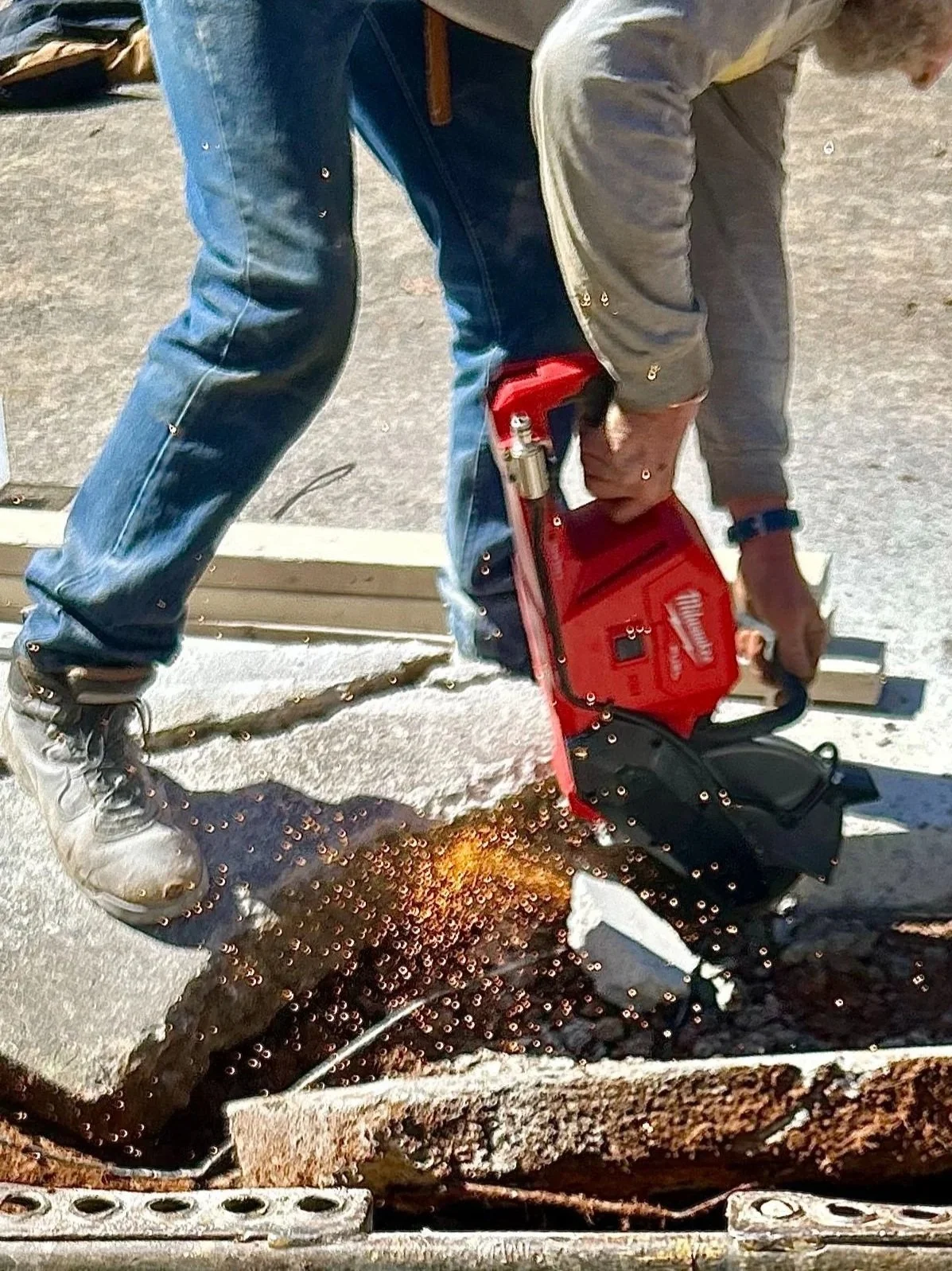 Construction worker wearing jeans and boots using a concrete cutter to cut through a large concrete slab. Sparks are visible from the cutting process.