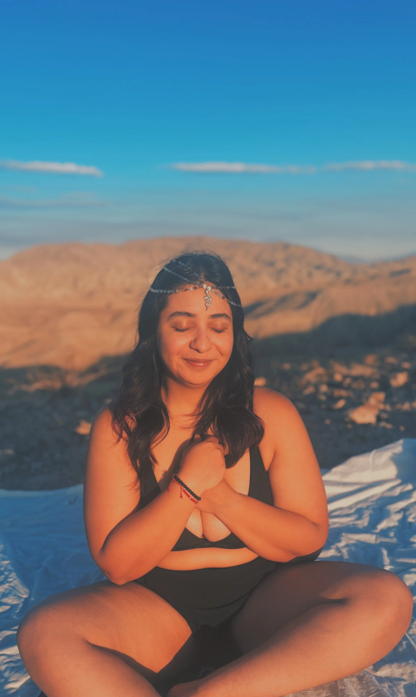 A woman sitting cross-legged on the ground outdoors, smiling with closed eyes, in a desert landscape during sunset, wearing black attire and jewelry.