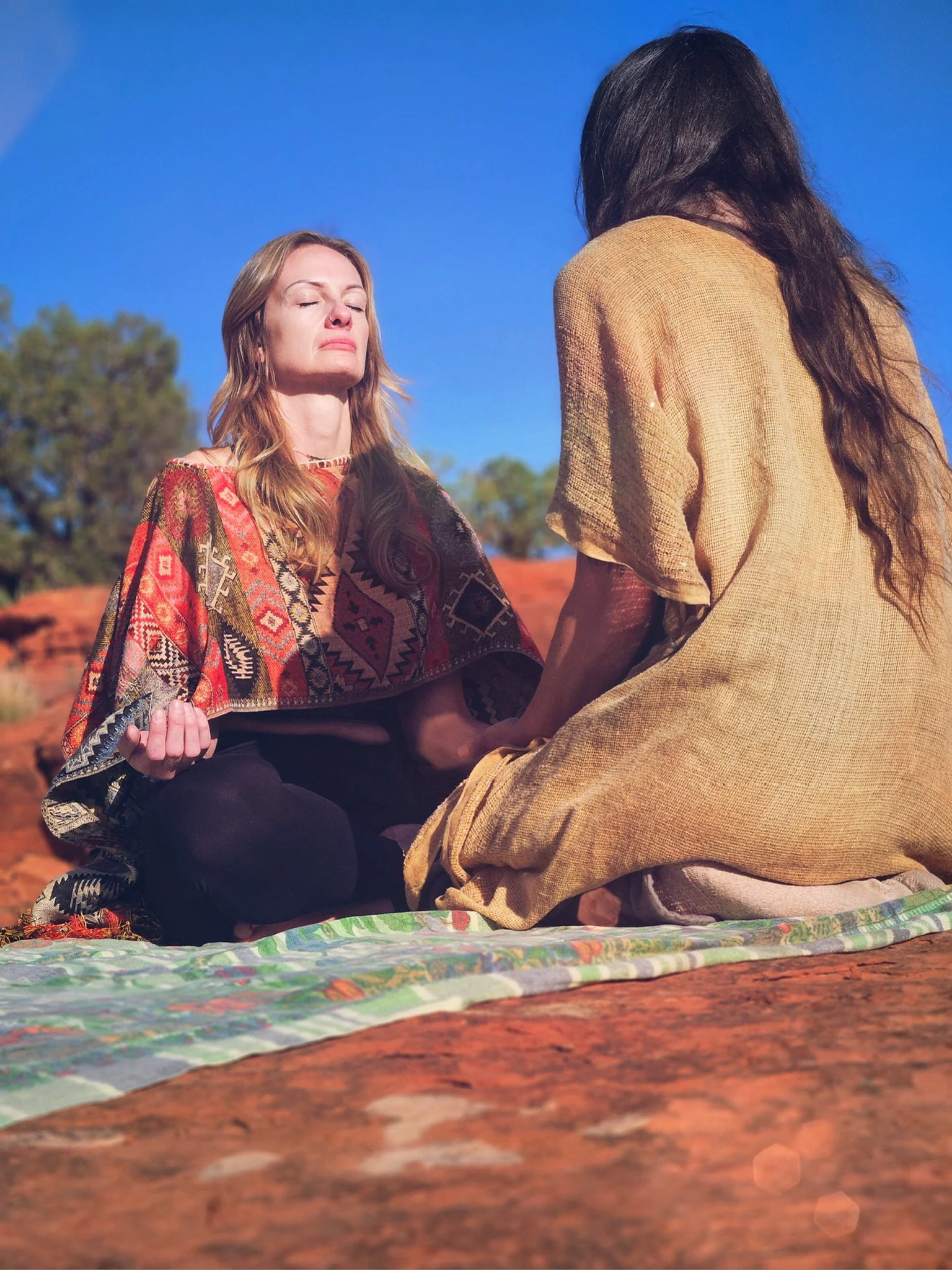 Two women sitting cross-legged on a colorful cloth outdoors, engaged in a spiritual or meditative practice during daytime with a clear blue sky and trees in the background.