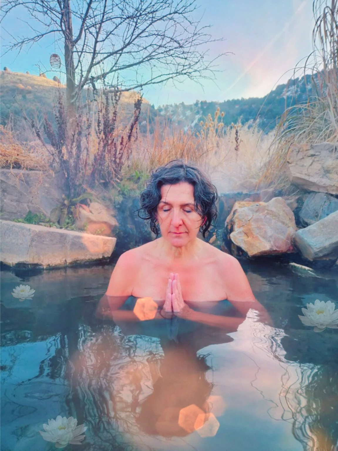 A woman with short curly dark hair practicing meditation in a natural hot spring surrounded by rocks and grasses, with mountains and clear sky in the background.