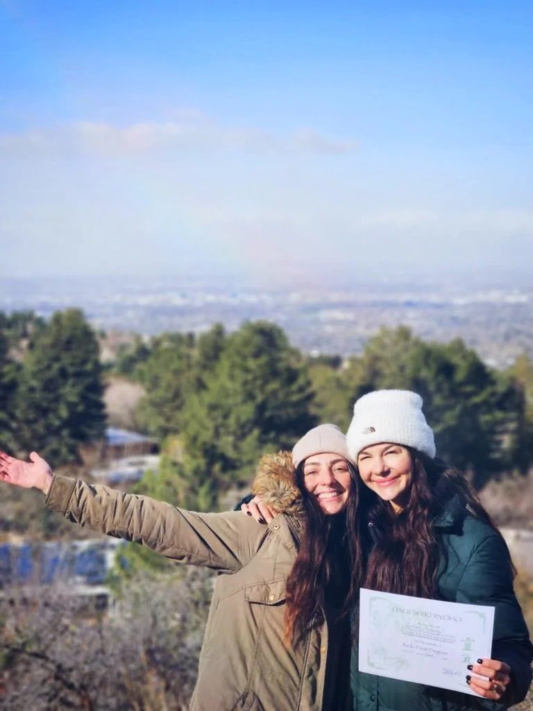Two smiling young women in winter jackets and beanies standing outdoors, one holding a certificate, with trees and a cityscape in the background.