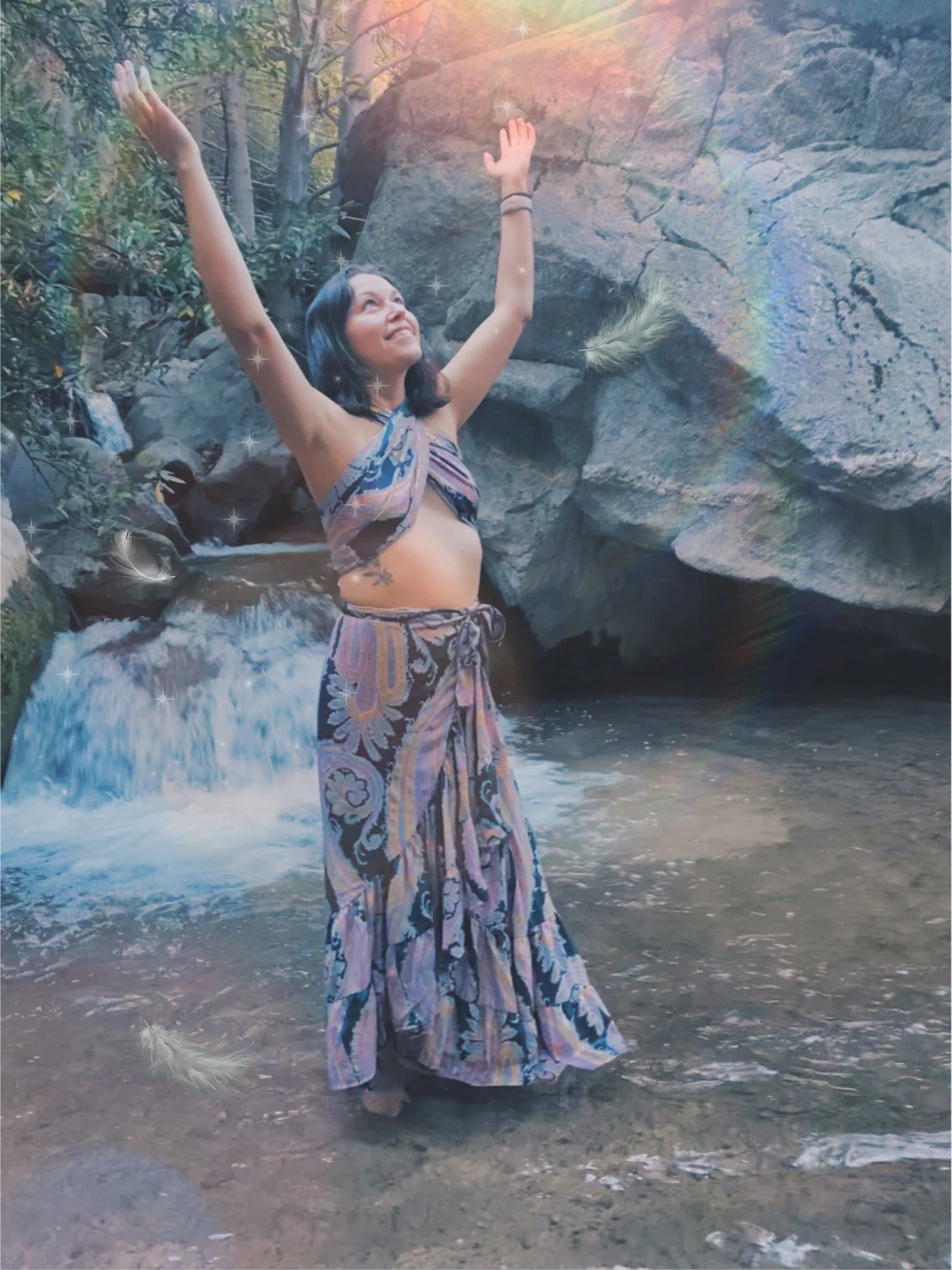 Woman in a colorful dress standing in a small stream near rocks, reaching upwards with arms outstretched, surrounded by nature, with sunlight shining through trees.