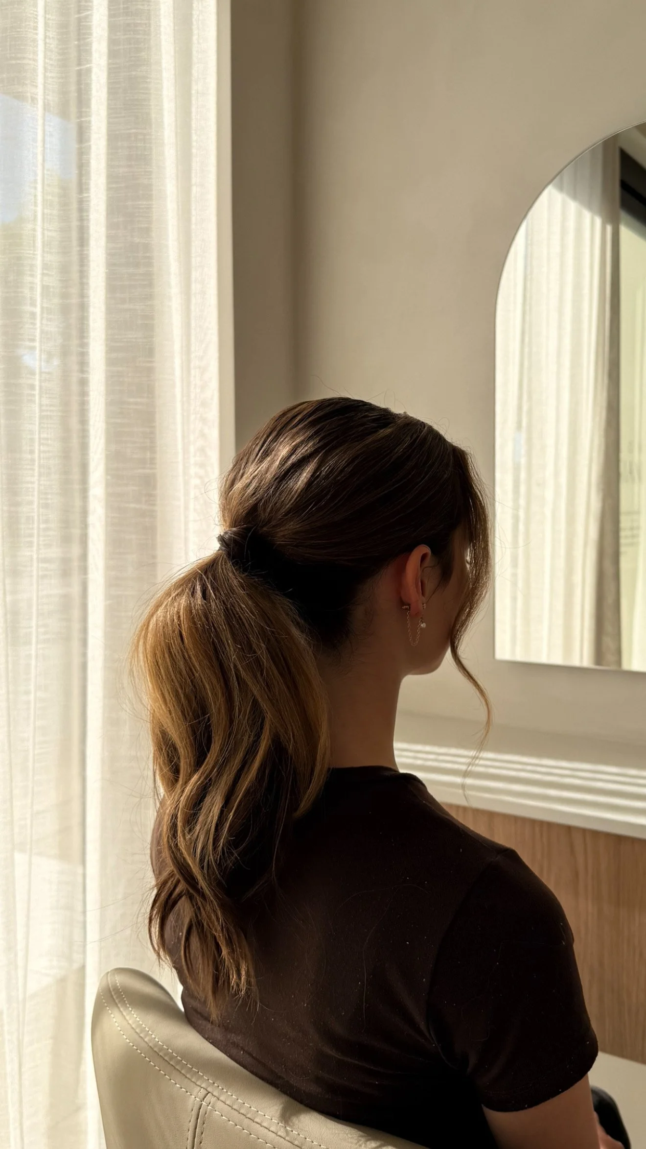 A woman with brown hair tied in a ponytail sits in a room, looking at a mirror. She is wearing earrings and a black shirt, with light coming through sheer curtains.