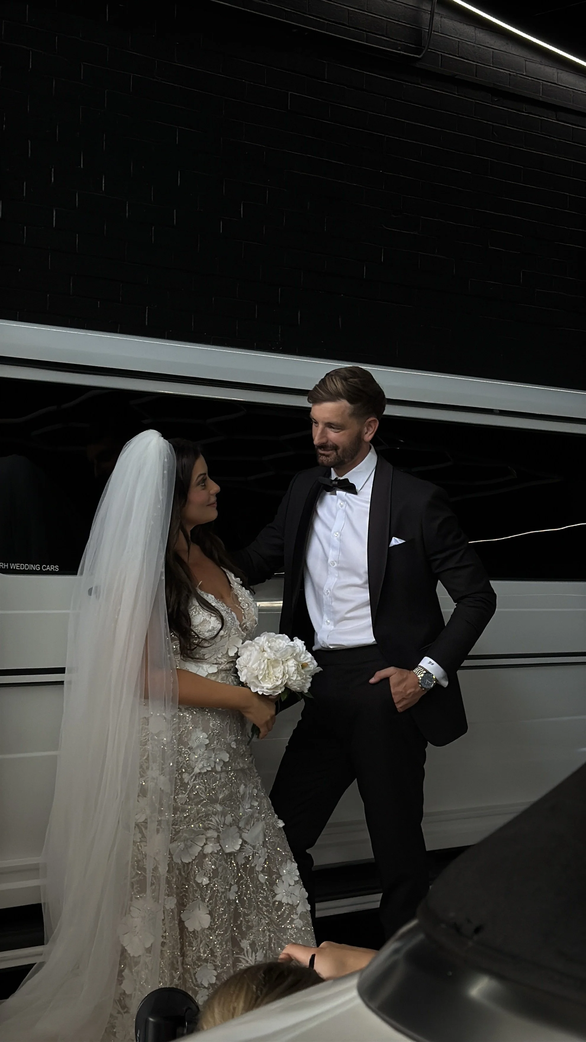 Bride and groom standing together in front of a white limousine, the bride holding a bouquet of white flowers, both dressed in formal wedding attire, sharing a moment.