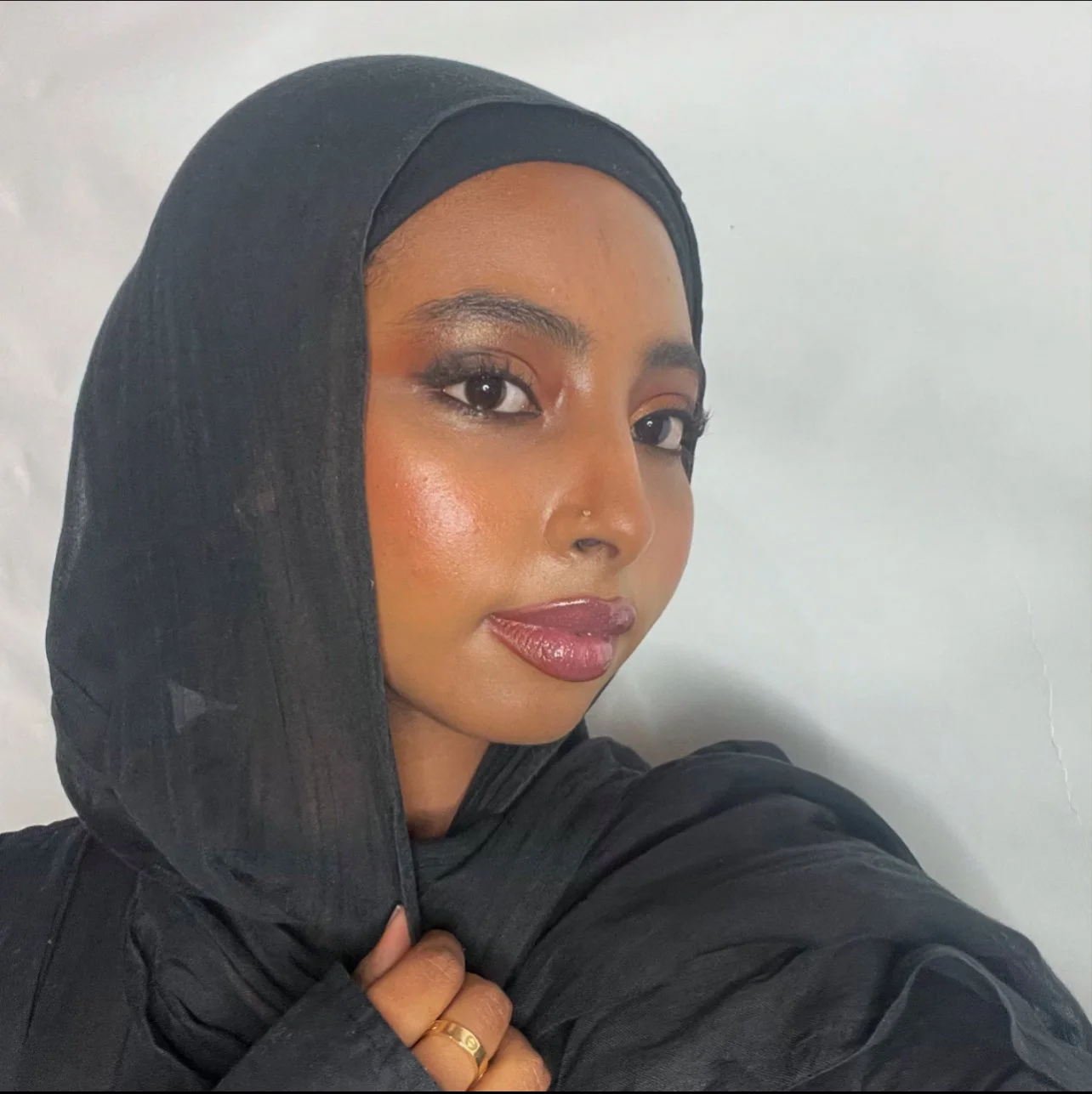 A woman with makeup, wearing a black headscarf, holding it with her left hand, against a plain light-colored background.