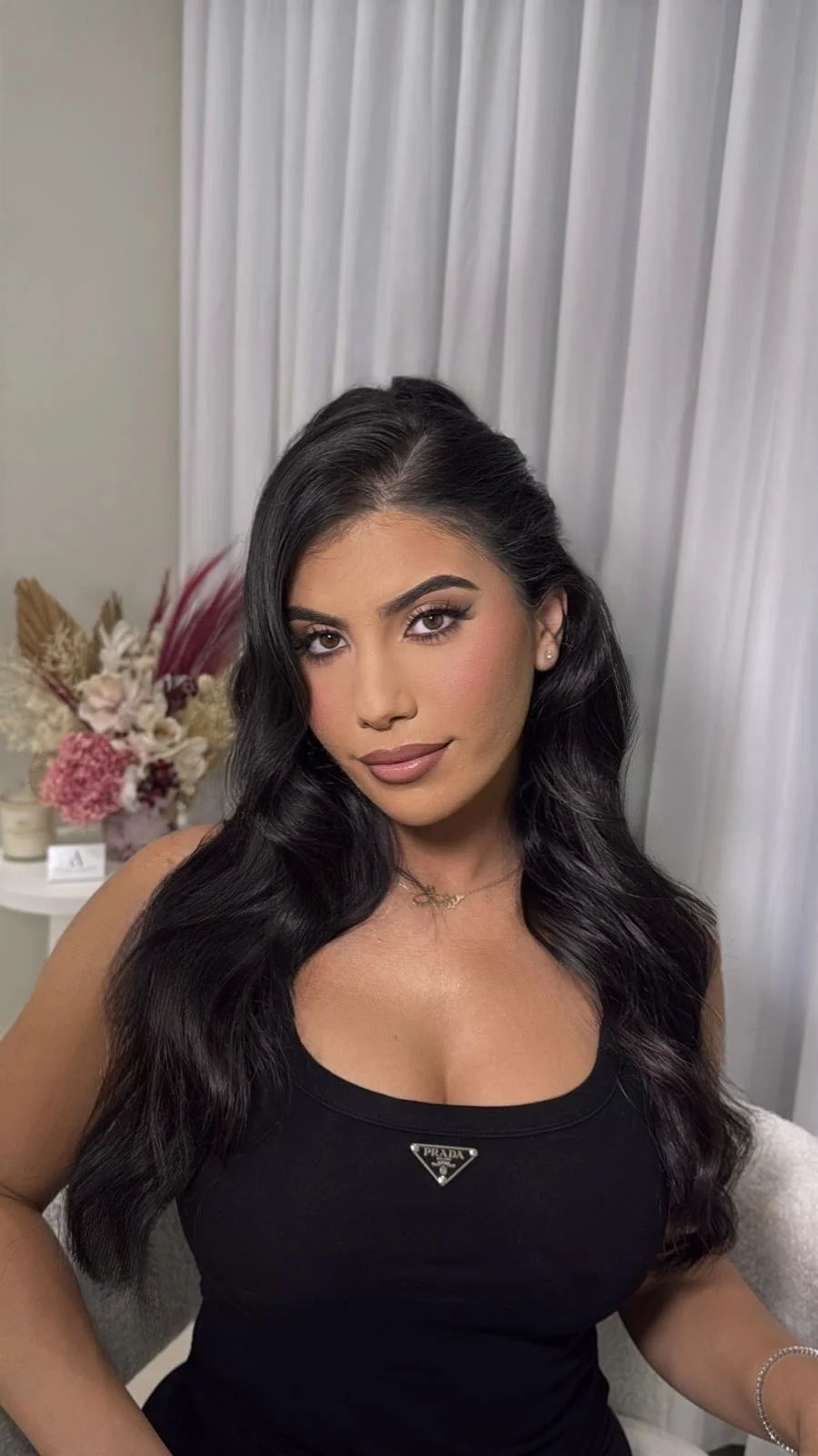 A woman with long, wavy black hair and makeup, wearing a black Prada tank top, sitting in front of white curtains with a floral arrangement in the background.