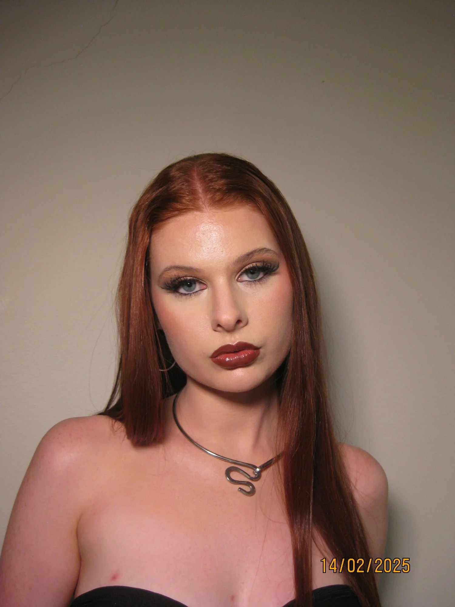A young woman with long red hair and fair skin is looking at the camera with a neutral expression. She is wearing makeup and a necklace with a swirl design. The background is plain and light-colored.