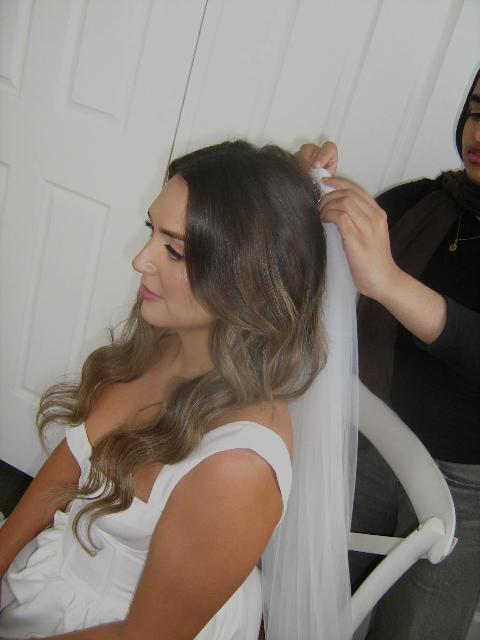 A woman with long wavy hair seated, having her hair styled or arranged by another woman.