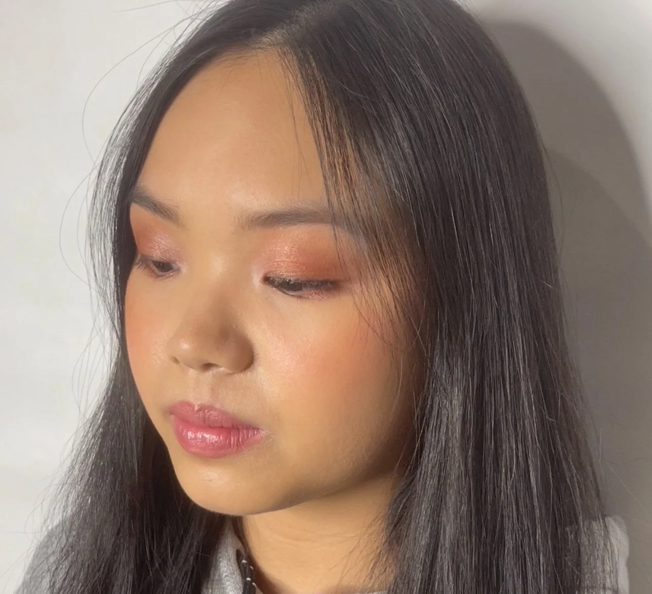 A young woman with long dark hair, wearing makeup with soft peach eyeshadow, pink lipstick, and a subtle blush, looking slightly downward.